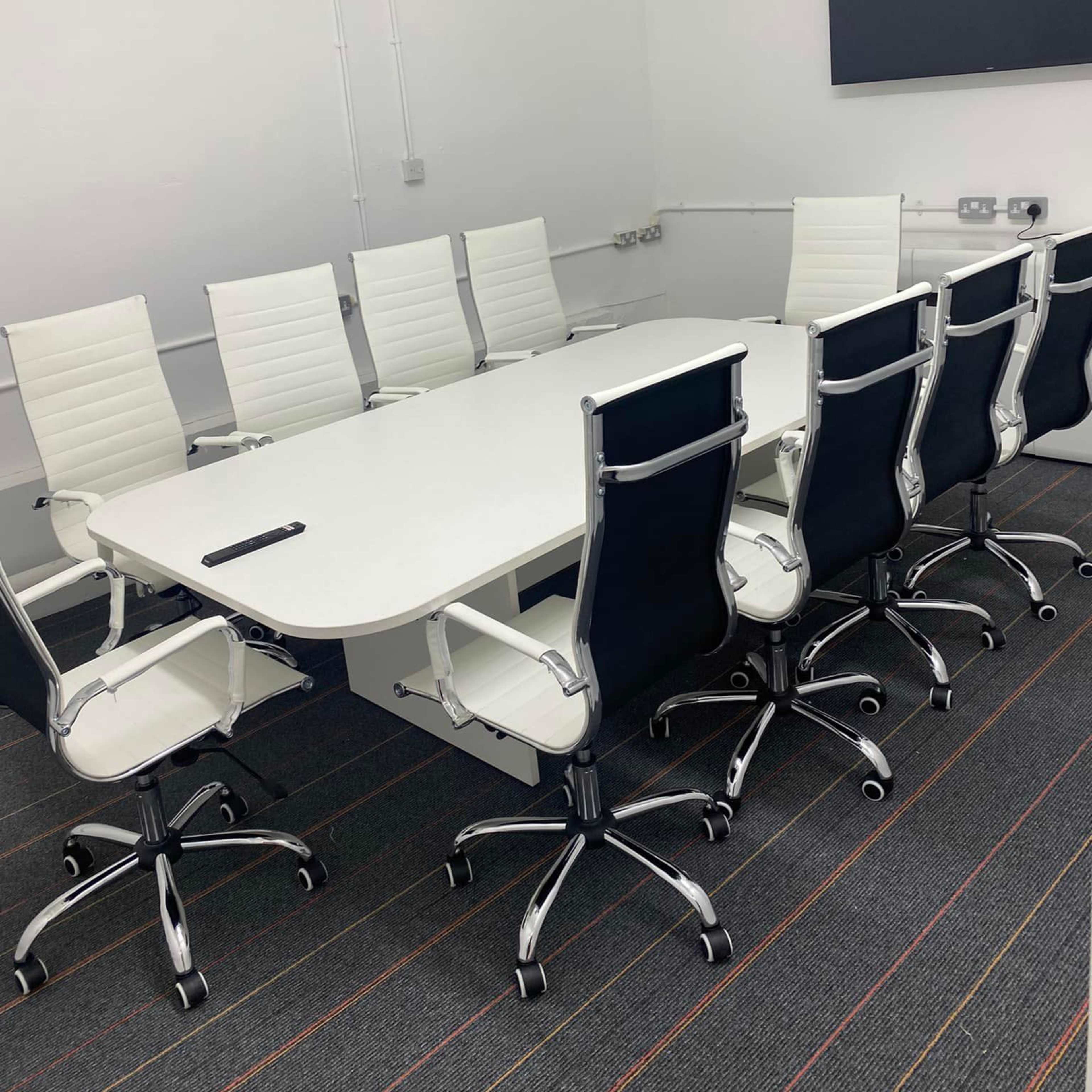 A modern conference room features a long white table surrounded by eight white and black office chairs, with a wall-mounted screen in the background.