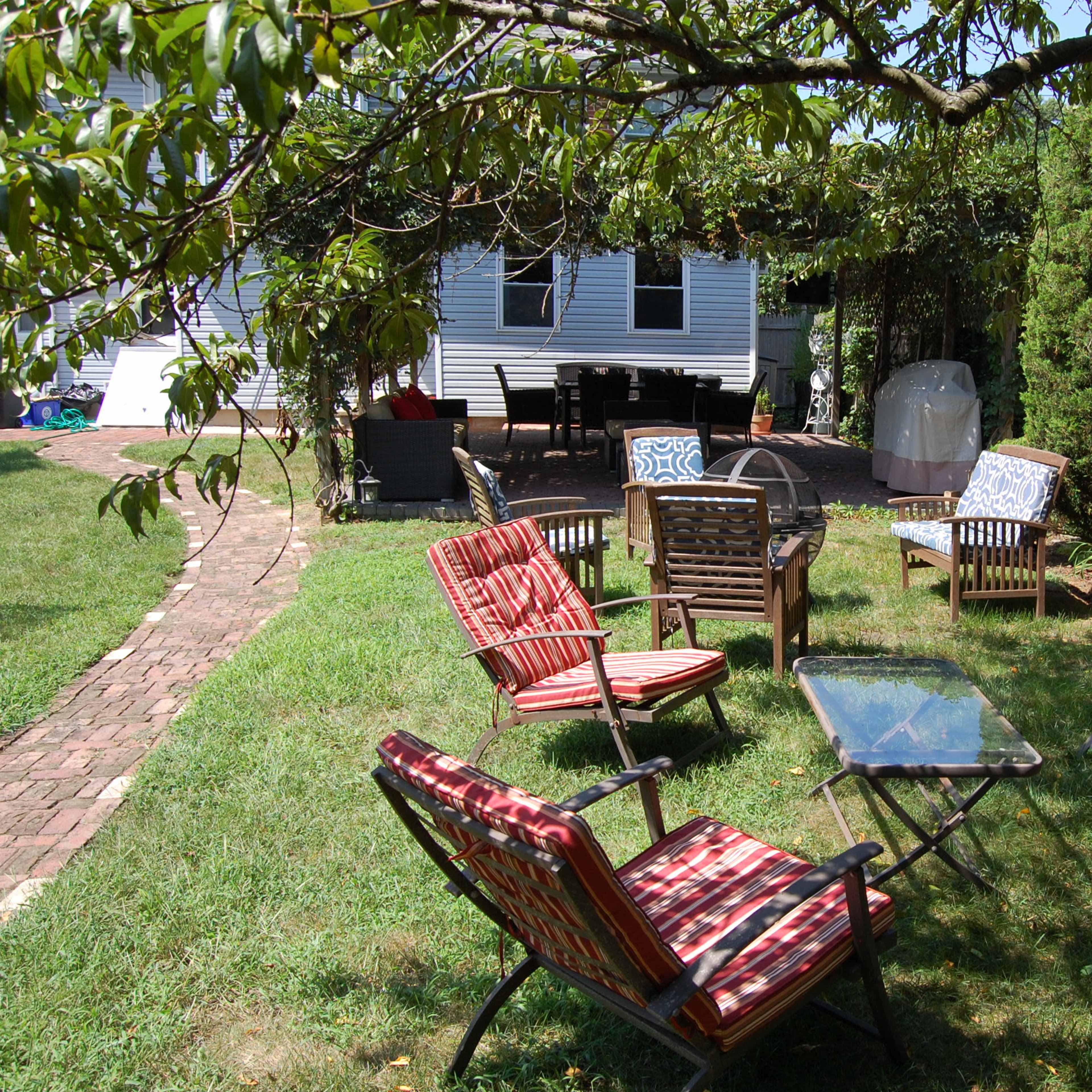 A garden area features several chairs with cushions arranged around a table, with a pathway leading to a house in the background.
