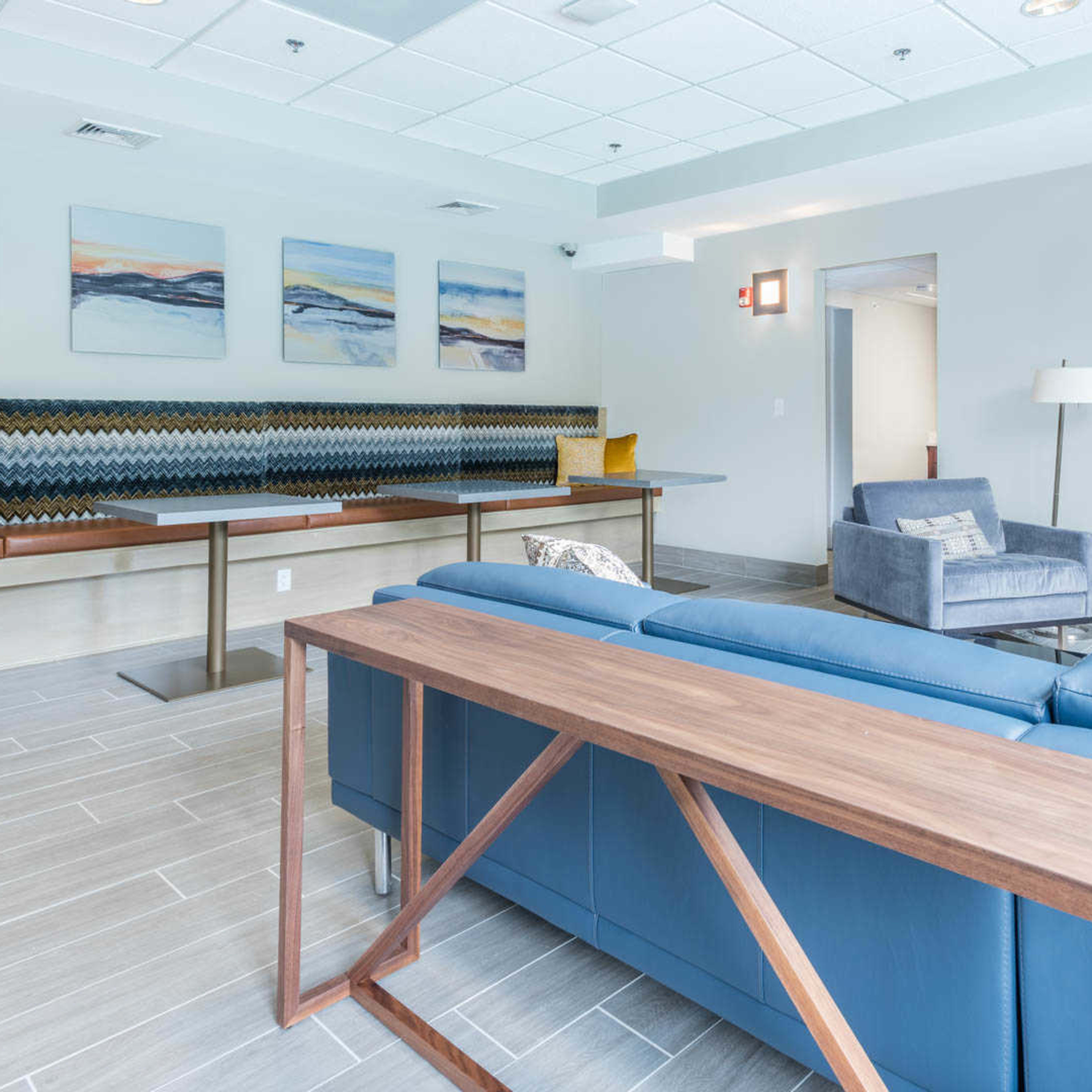 A modern lounge area with a blue sofa, a wooden table, an armchair, and abstract wall art.