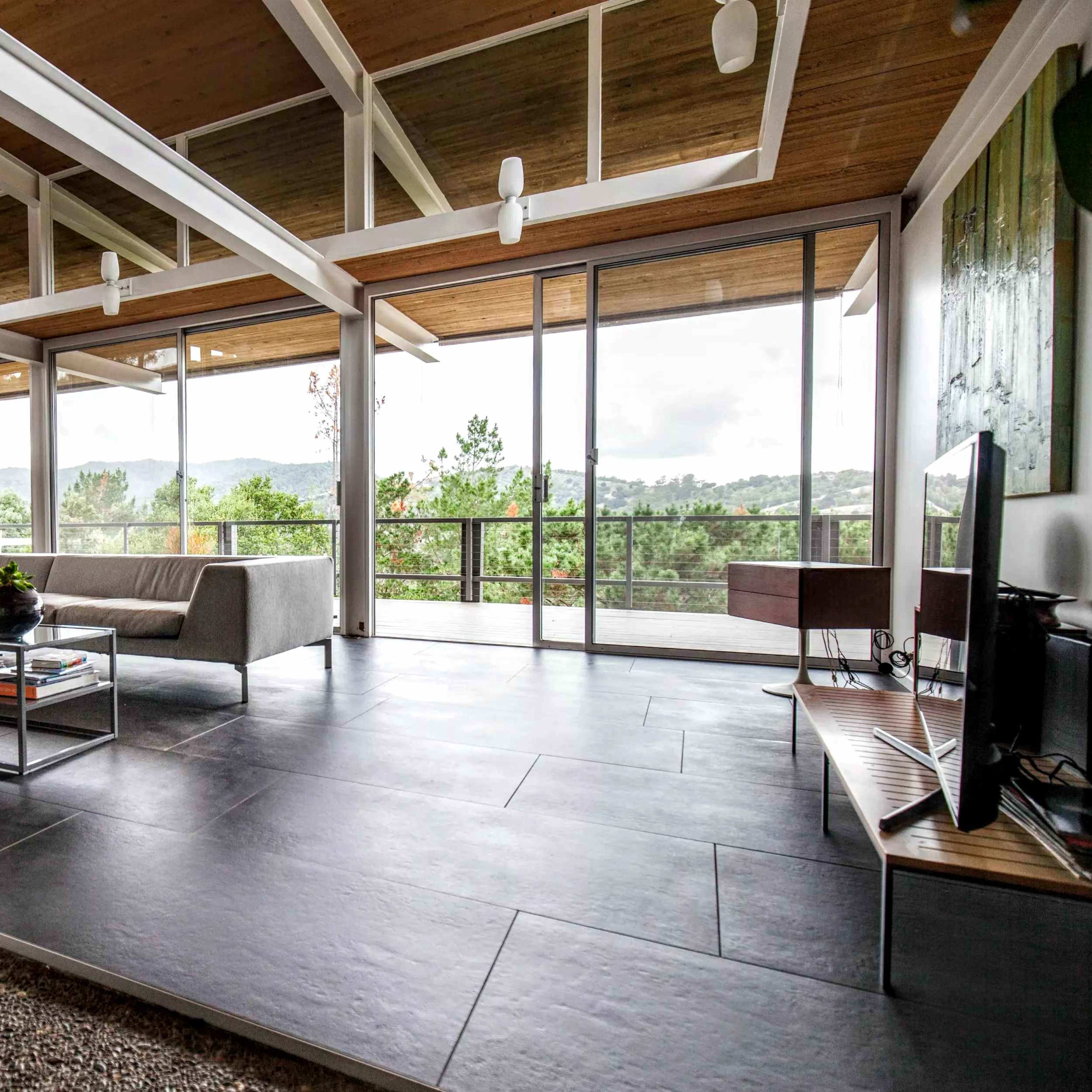 A modern living room with large windows, a gray sofa, a glass coffee table, and a view of a mountainous landscape.
