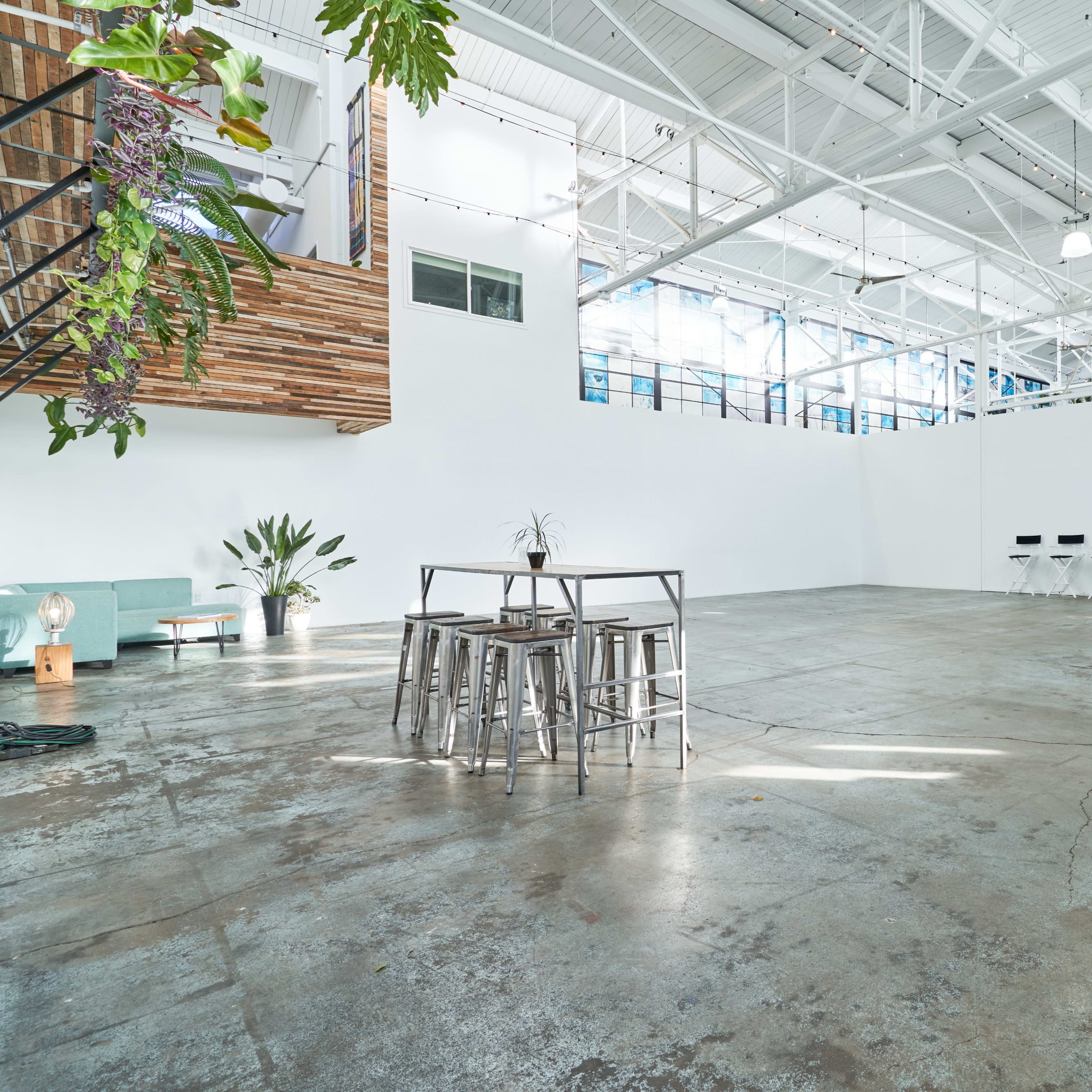 A spacious, empty industrial-style room features concrete flooring, large windows, and minimal furniture, including a metal table and stools.