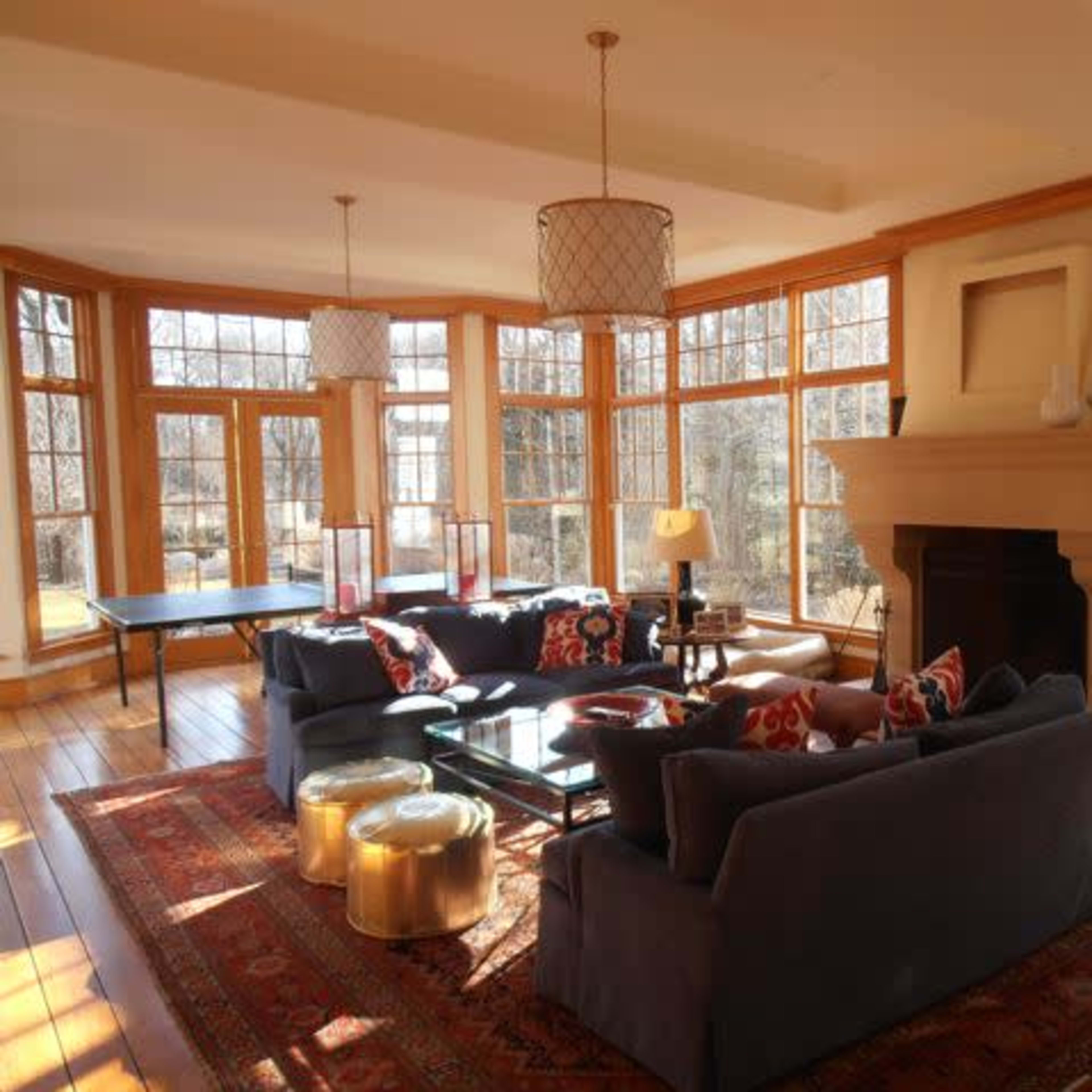 A bright living room features large windows, comfortable seating, and a central fireplace, with decorative tables and lamps.