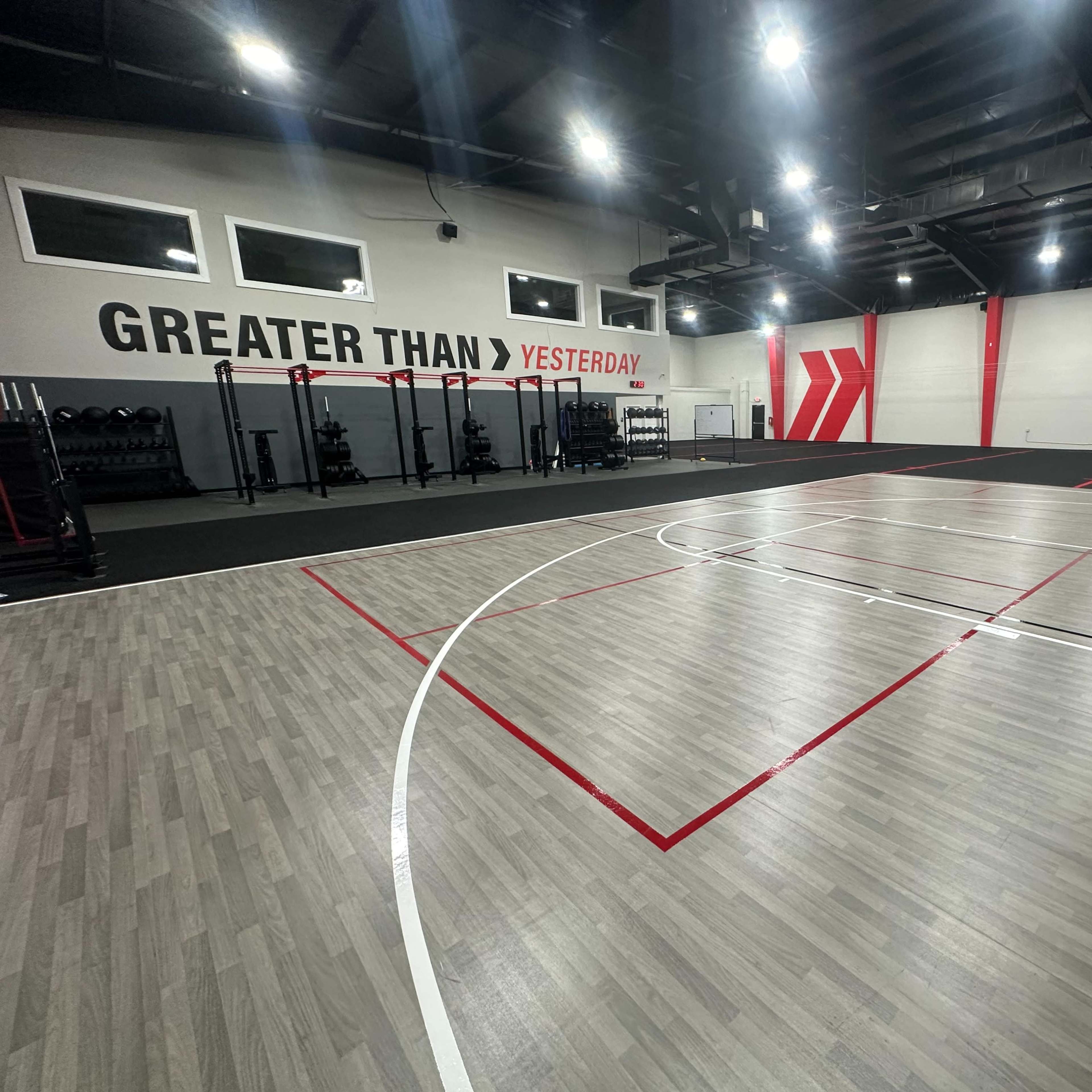 A modern indoor gym with a multi-use basketball court, workout equipment along the walls, and motivational signage on one side.