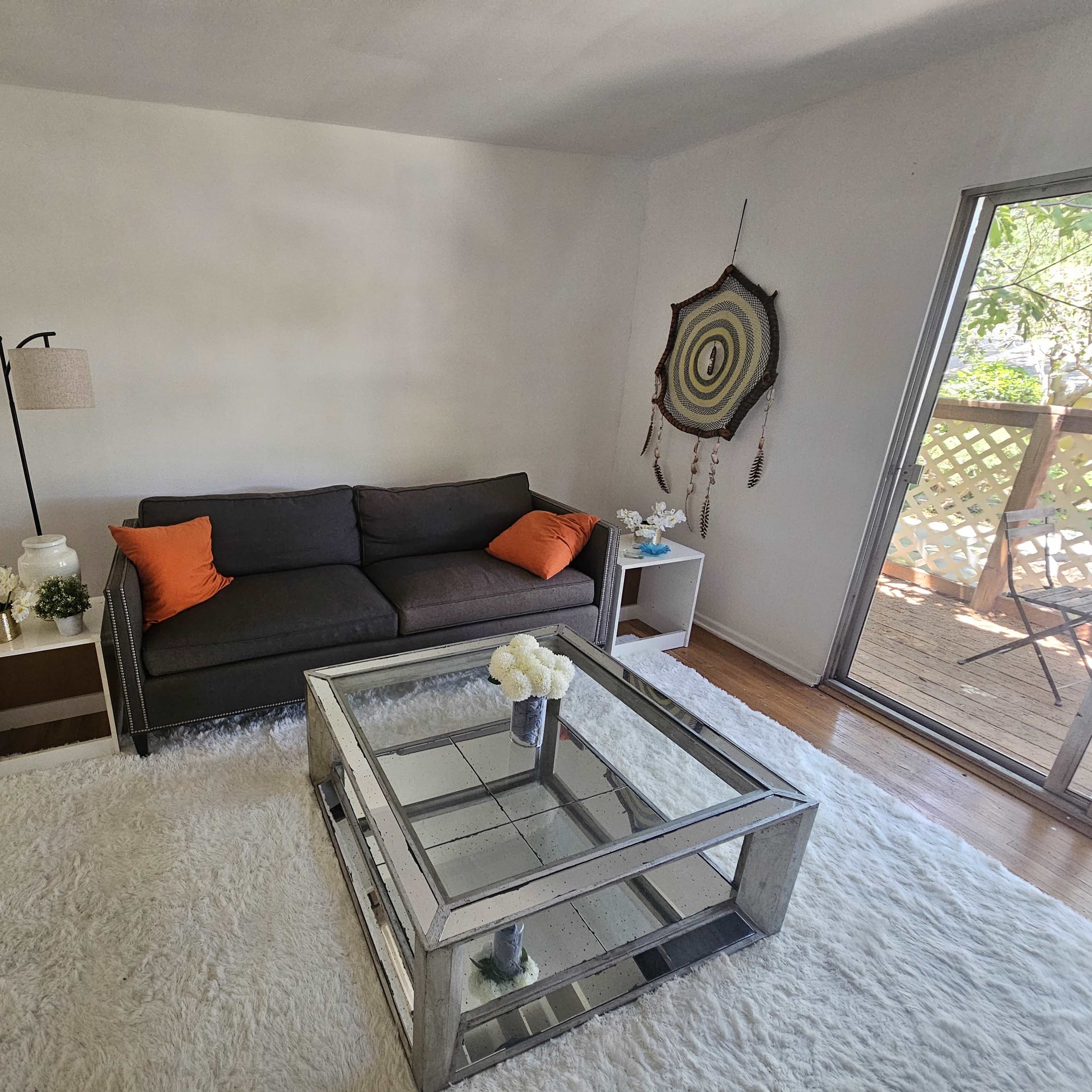 The room features a gray sofa with orange cushions, a glass coffee table, and a rug, alongside a lamp and decorative items, with a view of a wooden deck through the sliding door.