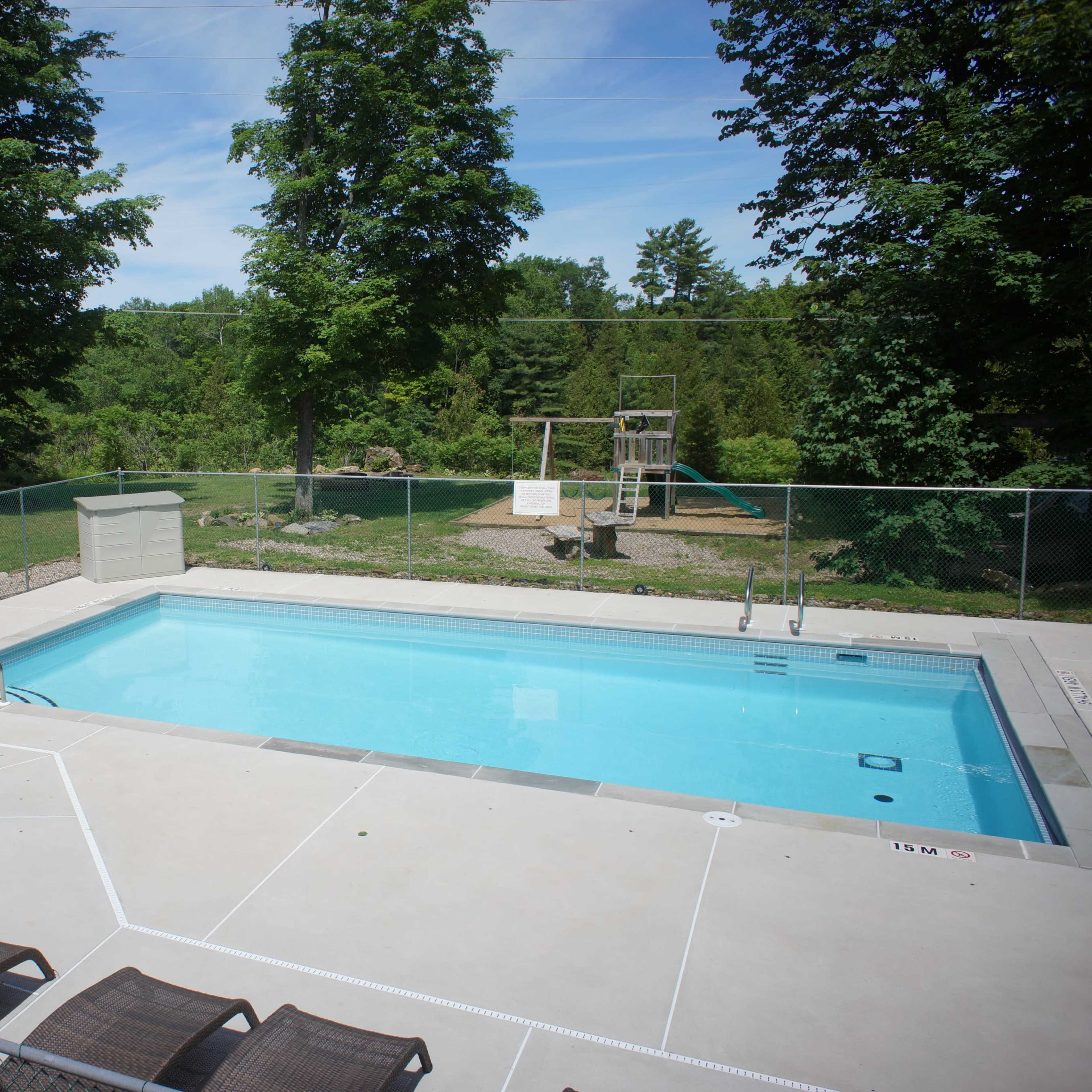 A rectangular swimming pool surrounded by a fenced area, with a hot tub and chairs nearby, and playground equipment in the background.