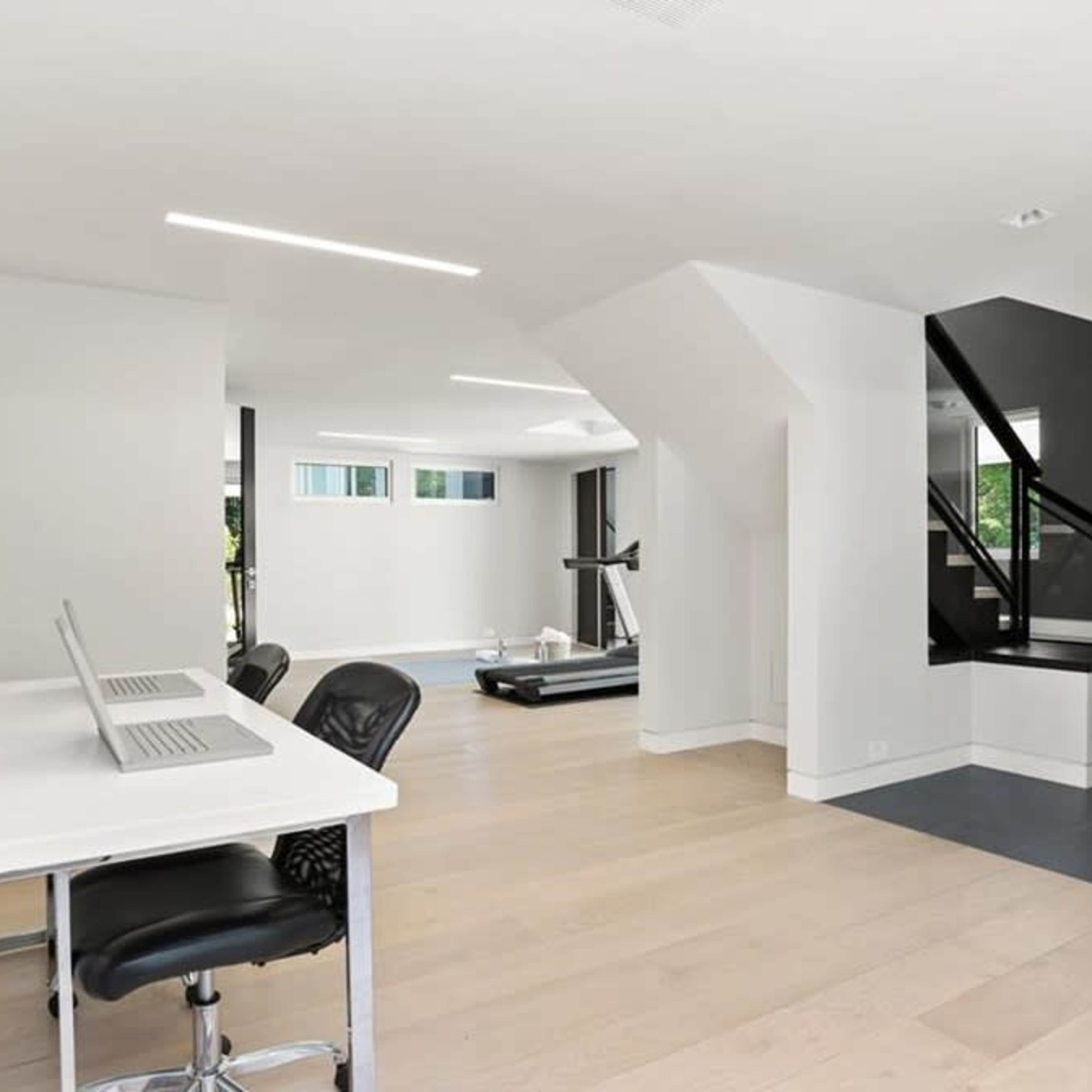 A modern interior space featuring a home office area with a desk and chair, a treadmill in the background, and a staircase leading to an upper level.