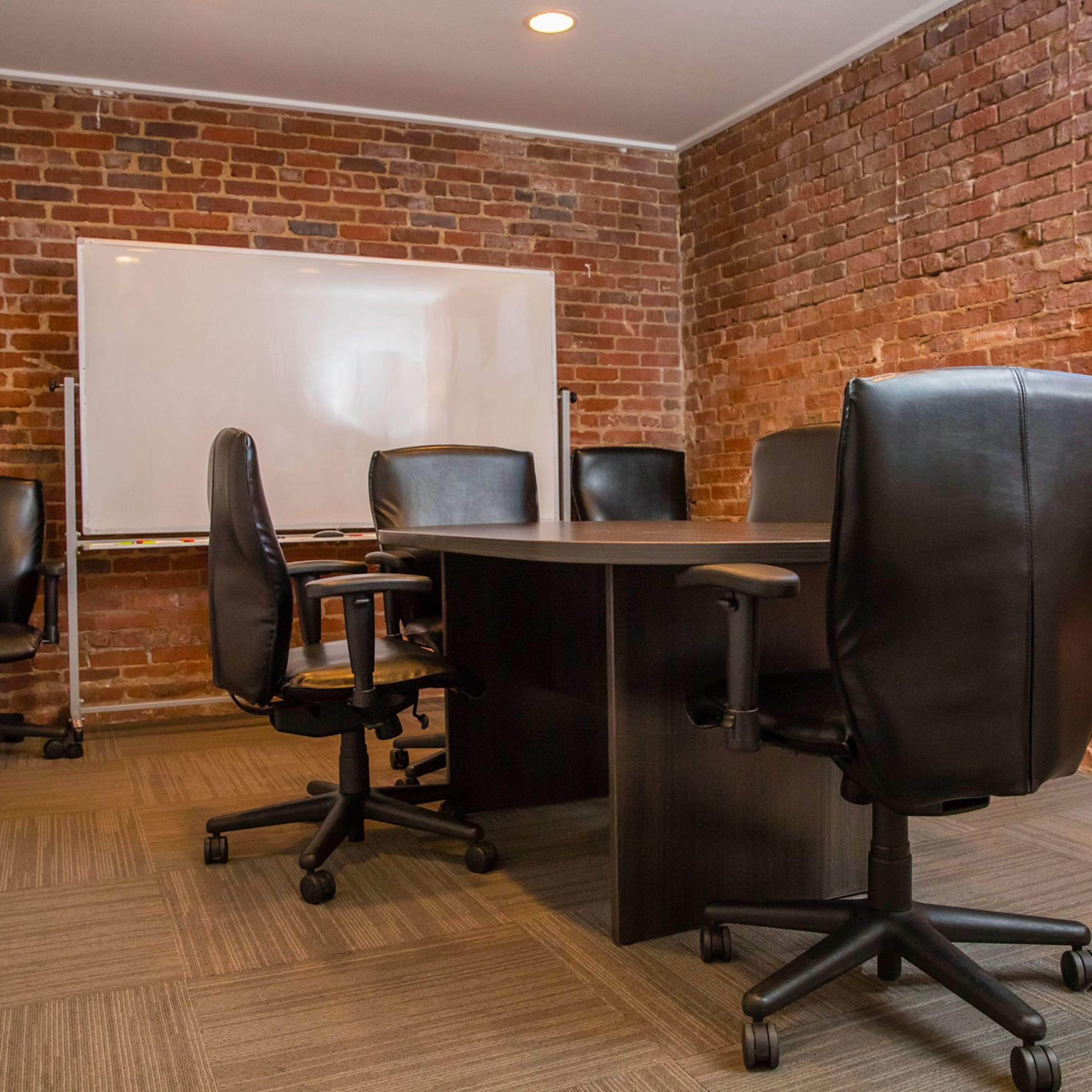 A meeting room features a large dark table surrounded by five black swivel chairs, with exposed brick walls and a whiteboard on a stand.