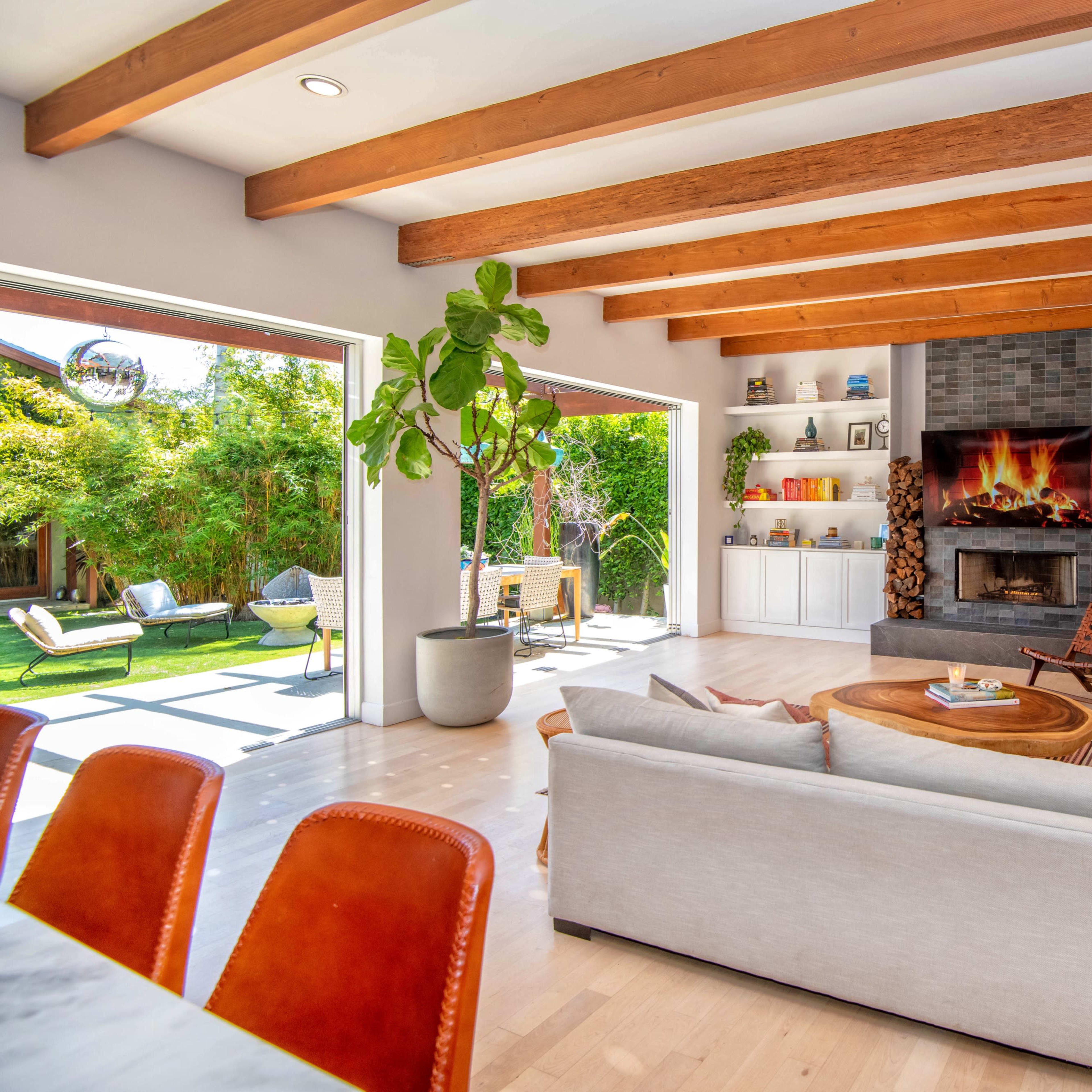 A modern living room features a light-colored sofa, a circular wooden coffee table, and a stone fireplace, with large openings leading to an outdoor patio and garden.