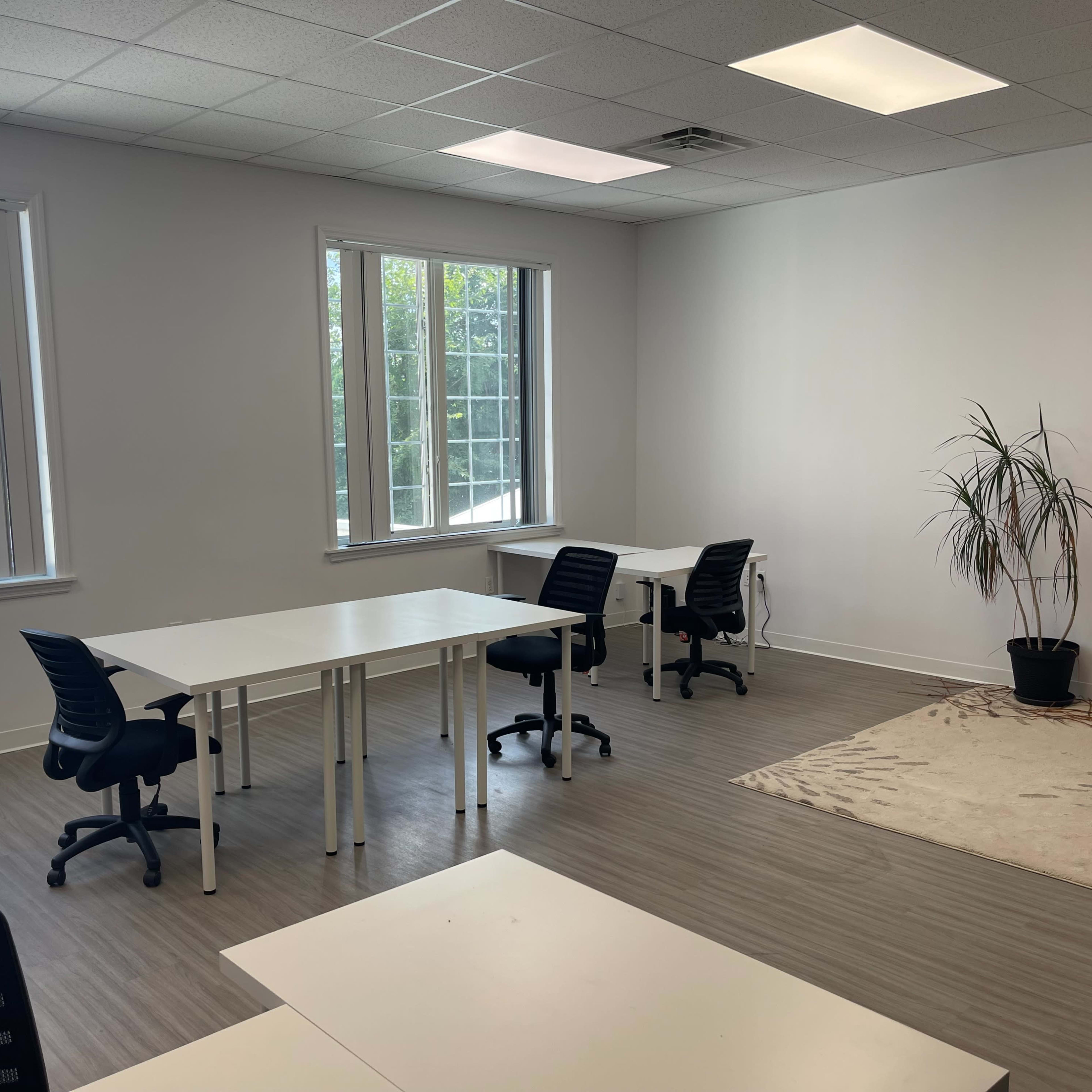 A bright, empty office space with several white tables and black chairs arranged around a light-colored rug and a potted plant in one corner.