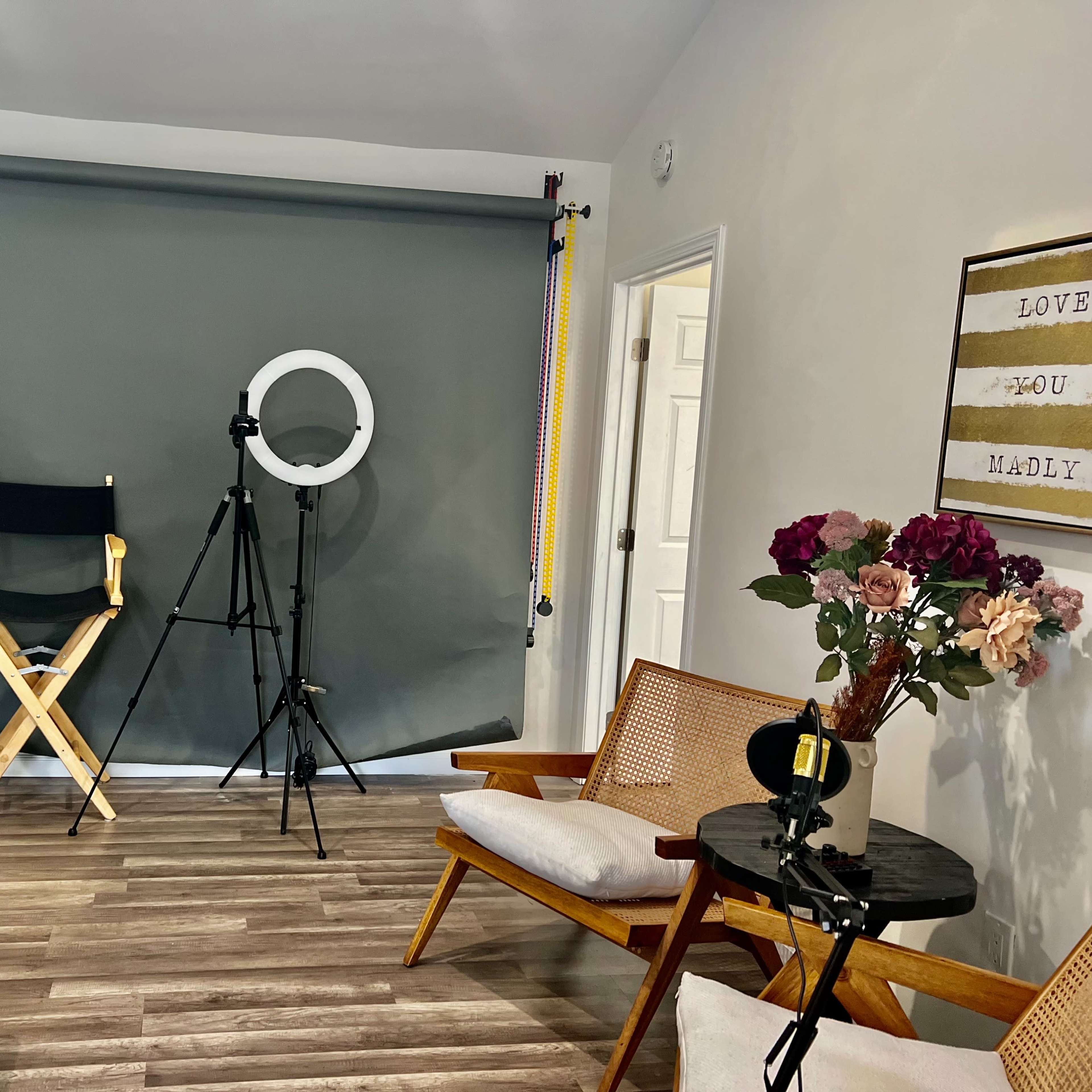 A well-lit room featuring a green backdrop, a director's chair, a ring light on a tripod, and two wooden chairs beside a small table with a vase of flowers.