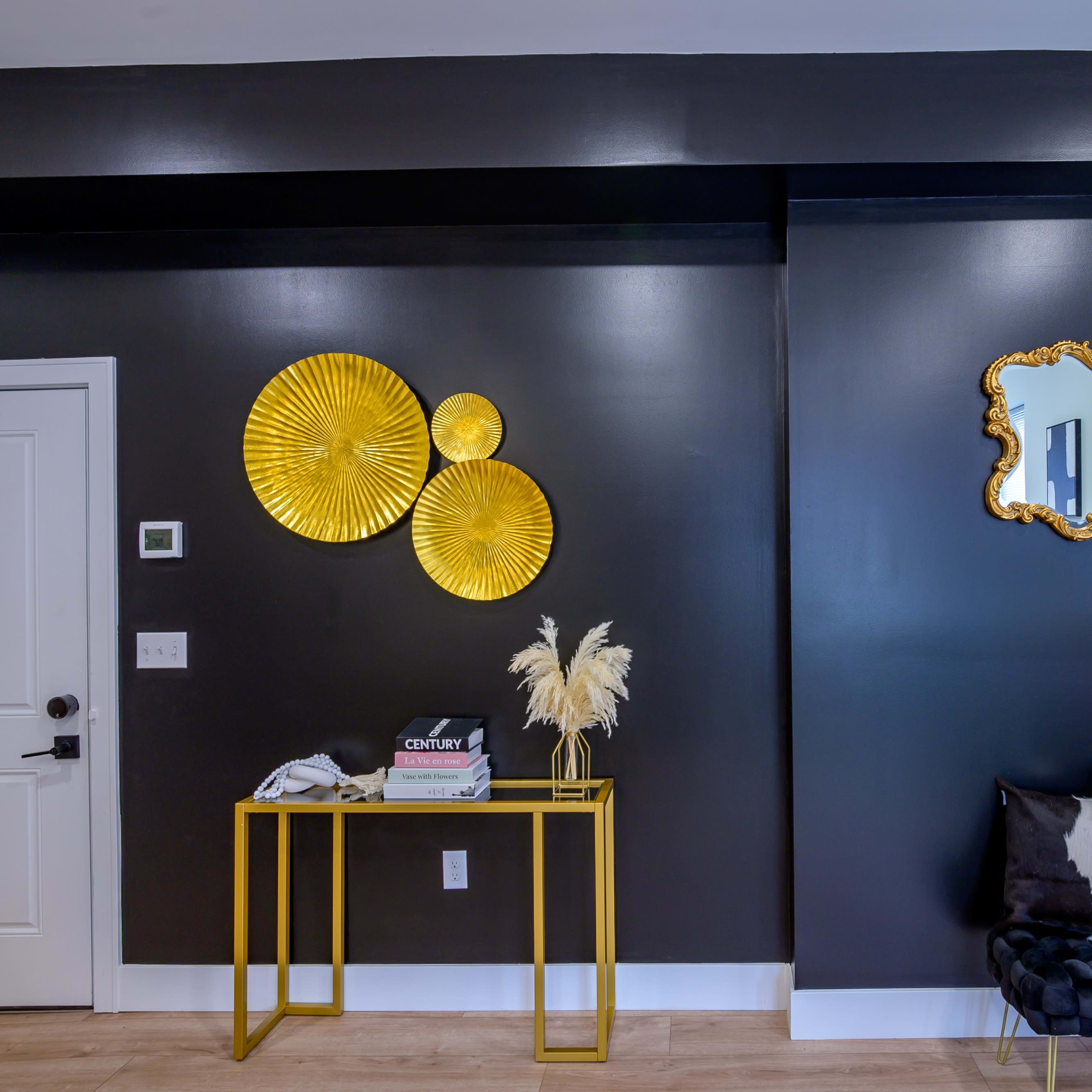 A modern entryway with a dark wall, three gold decorative circular wall pieces, a gold-framed mirror, a black and white patterned couch, and a gold side table displaying books and decorative items.