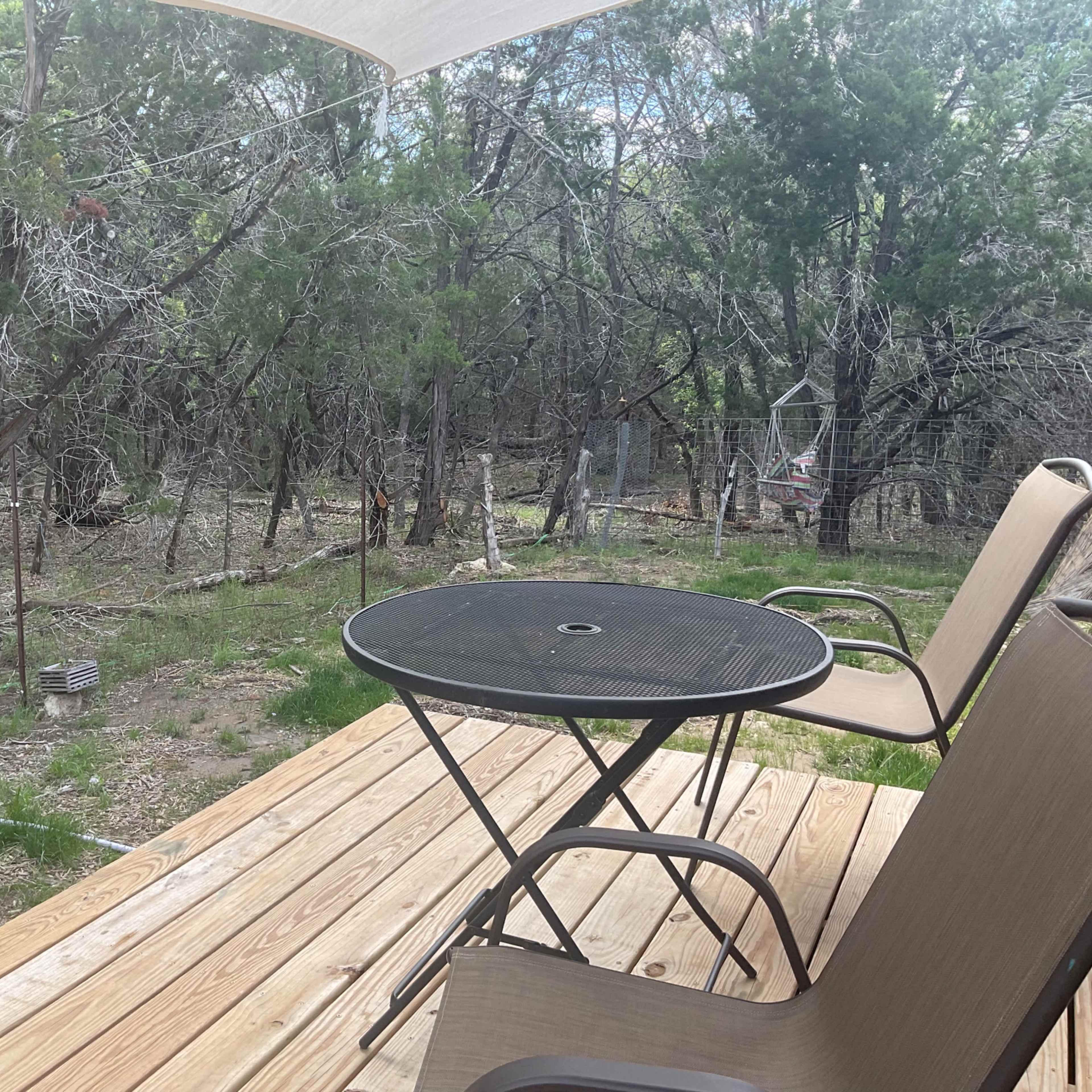 A wooden deck with two chairs and a small table overlooks a wooded area with sparse trees and underbrush.