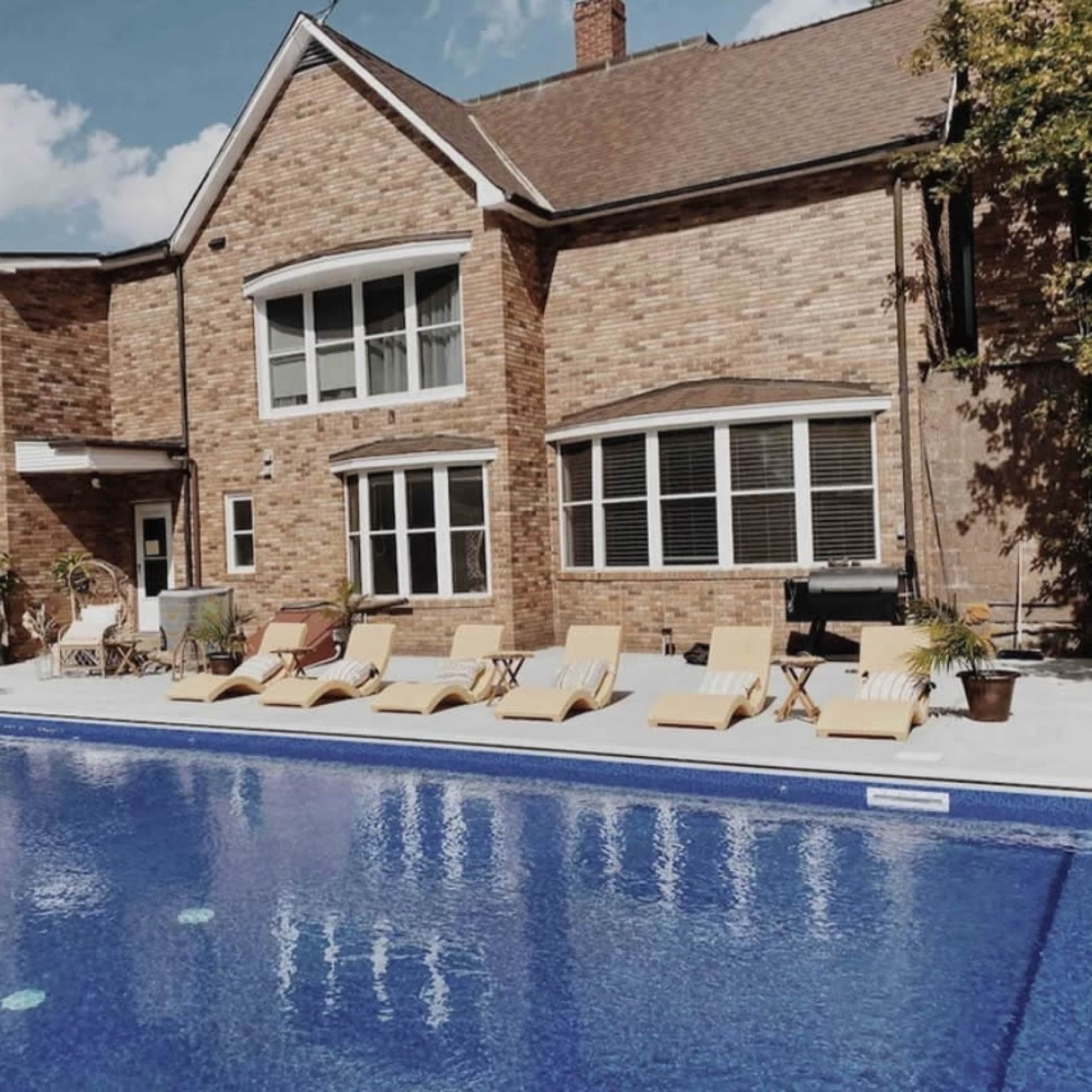 A brick house with large windows sits beside a blue swimming pool surrounded by lounge chairs and greenery.