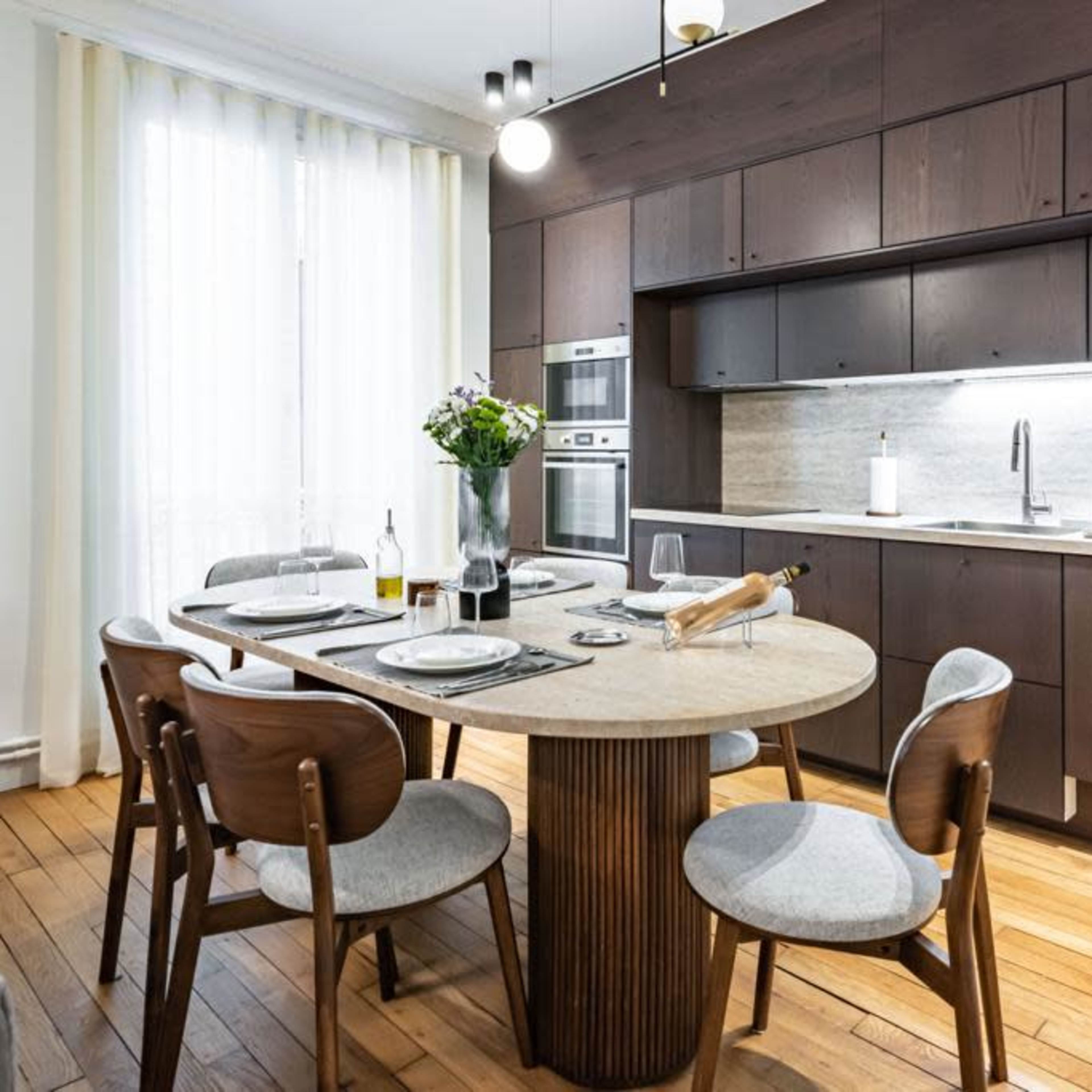 A modern kitchen features a round dining table surrounded by wooden chairs, with a countertop and cabinets in dark tones and large windows letting in ample light.
