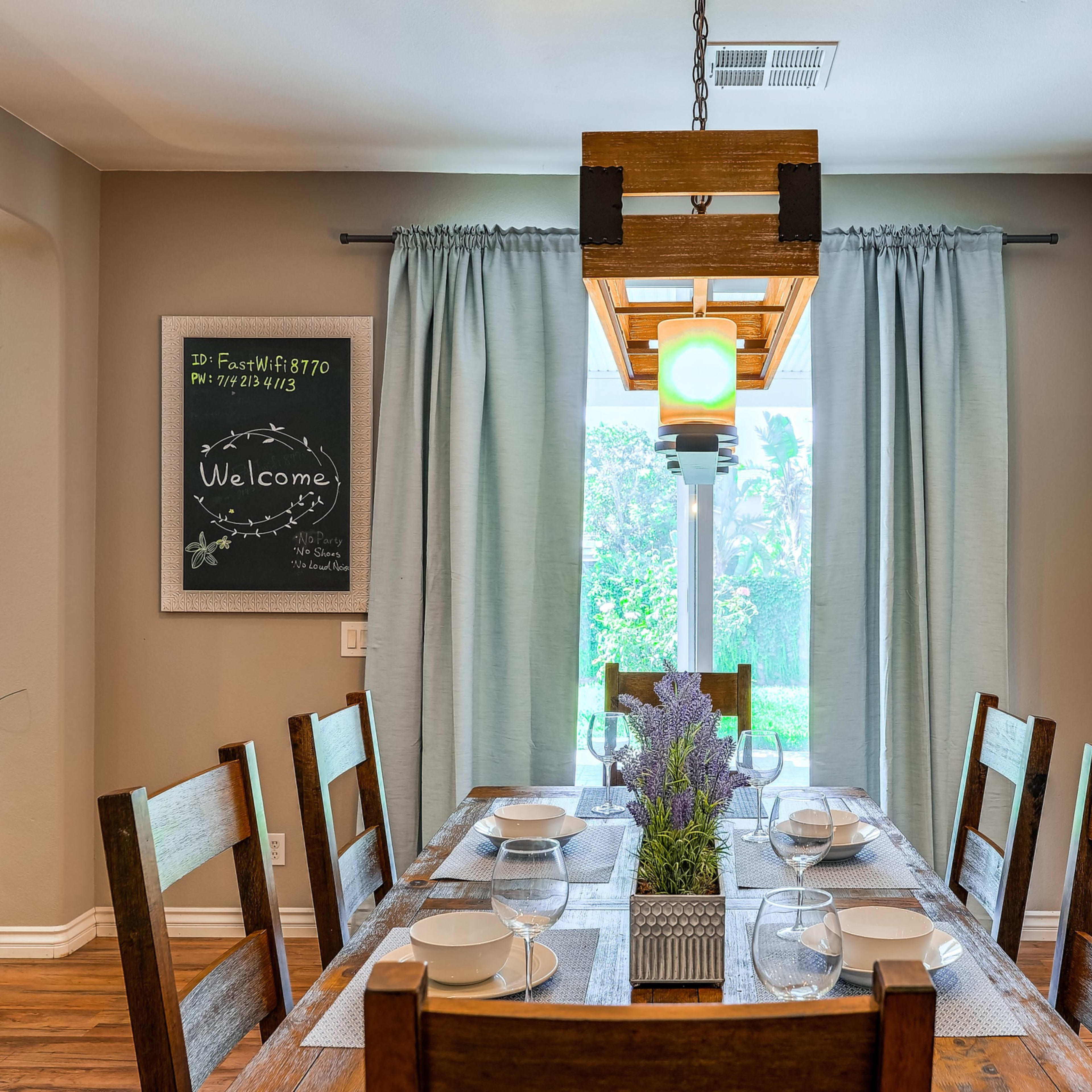 A wooden dining table is set for a meal, with plates arranged on it, positioned near a window with light green curtains and a chalkboard displaying a welcome message.