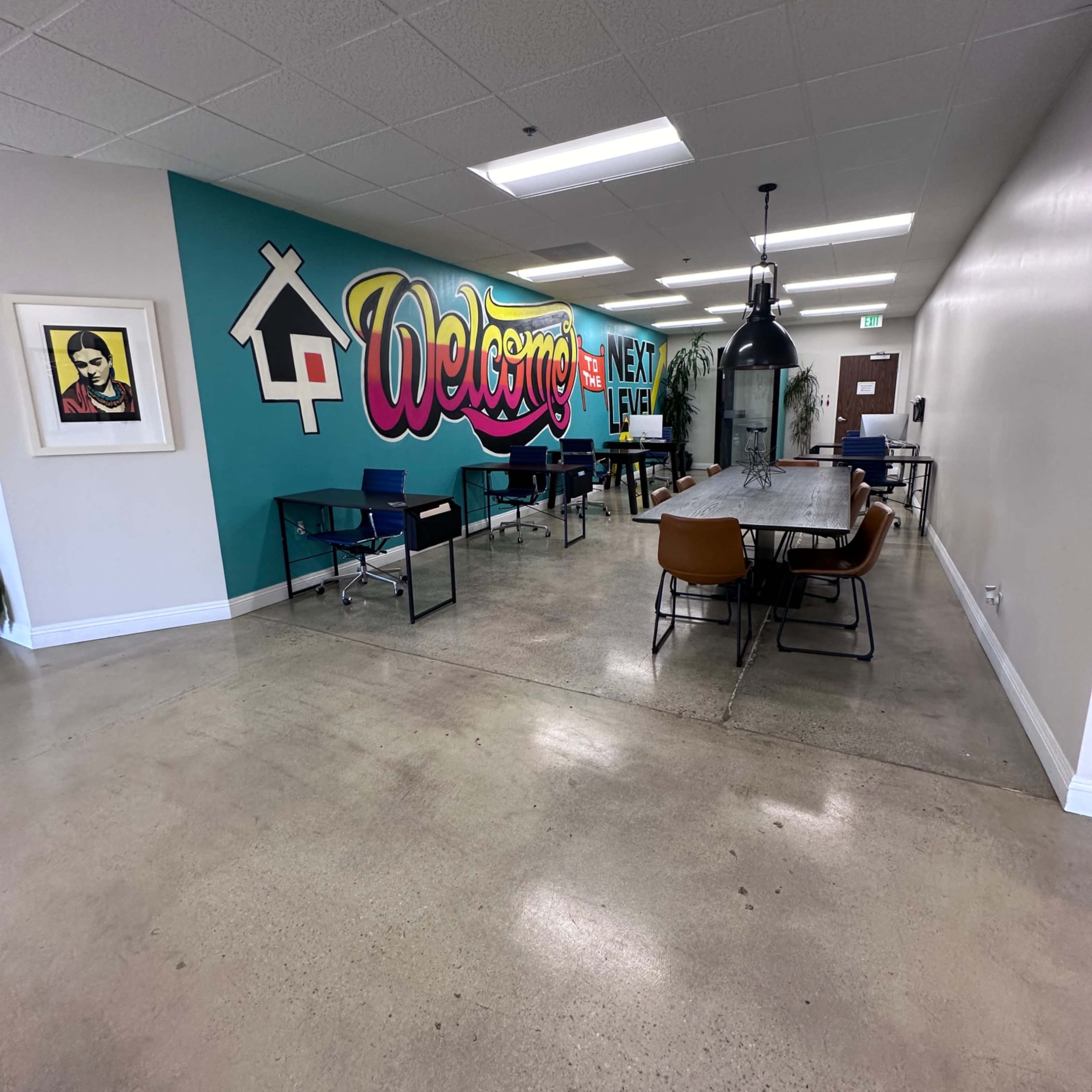 A spacious office area featuring a colorful "Welcome" mural, several desks with chairs, and a large communal table in a well-lit environment.