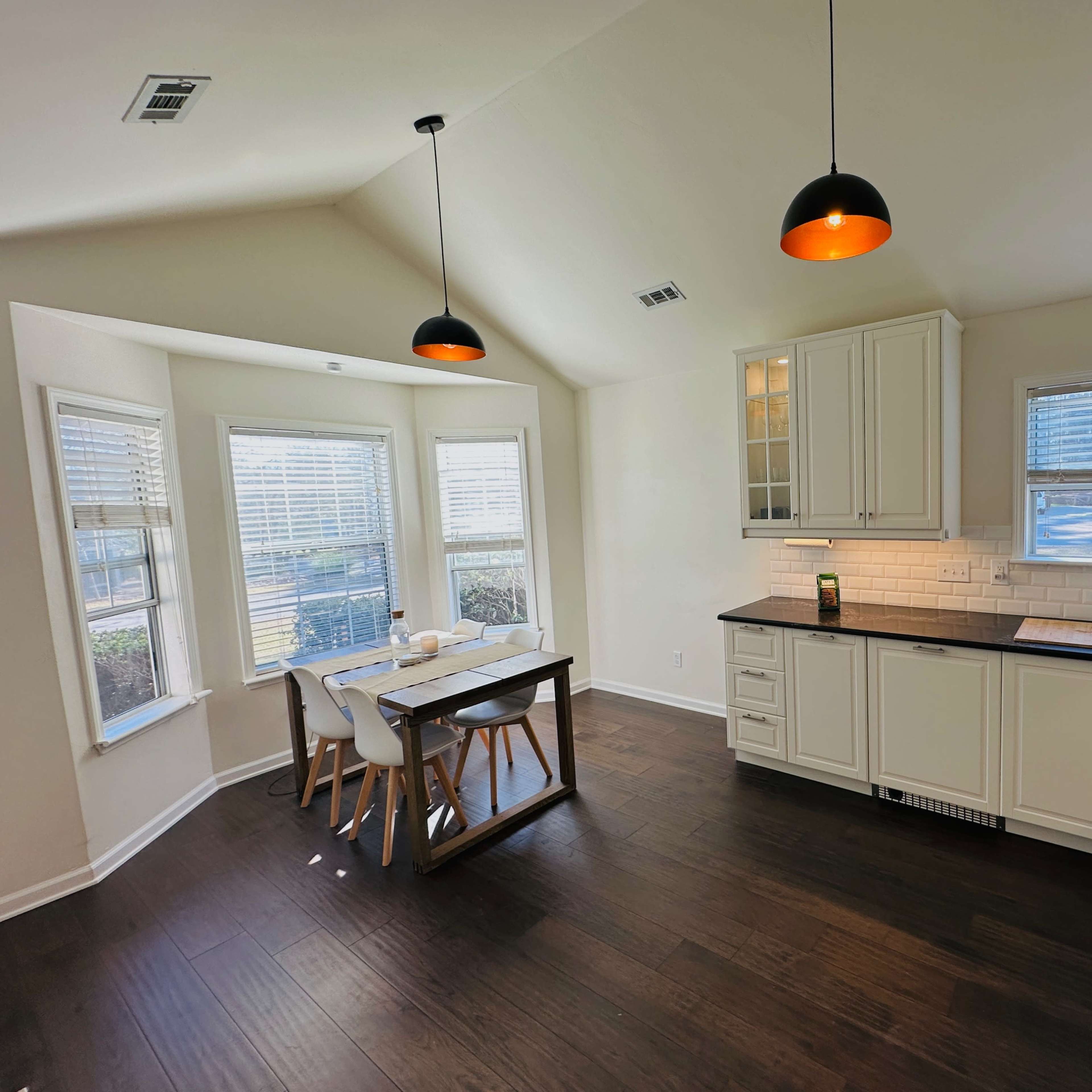 A modern kitchen features white cabinetry, a dark countertop, a dining table with four chairs, and large windows allowing natural light.