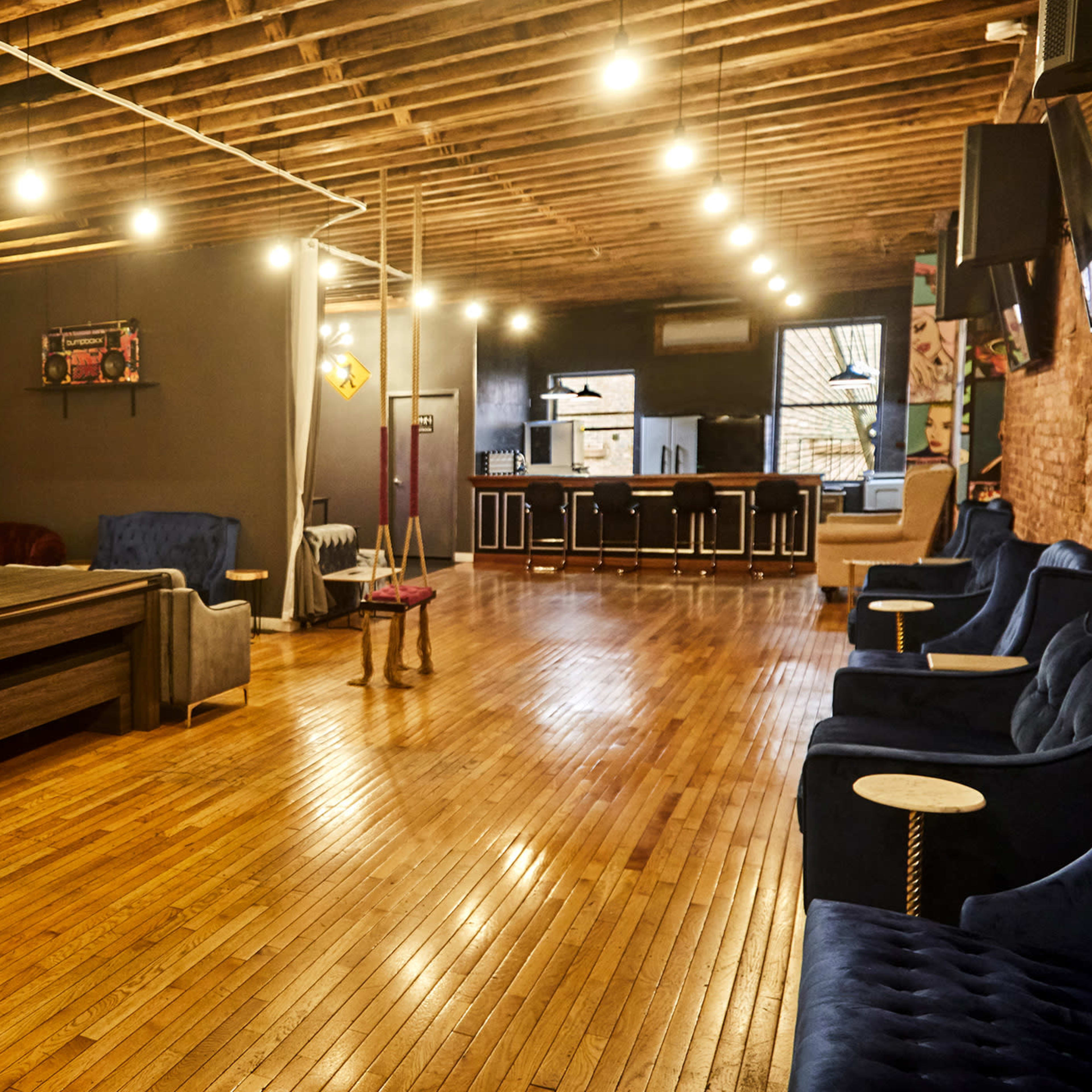 A spacious indoor area with wooden flooring, featuring various seating arrangements, including sofas and swings, and a bar counter at the far end.