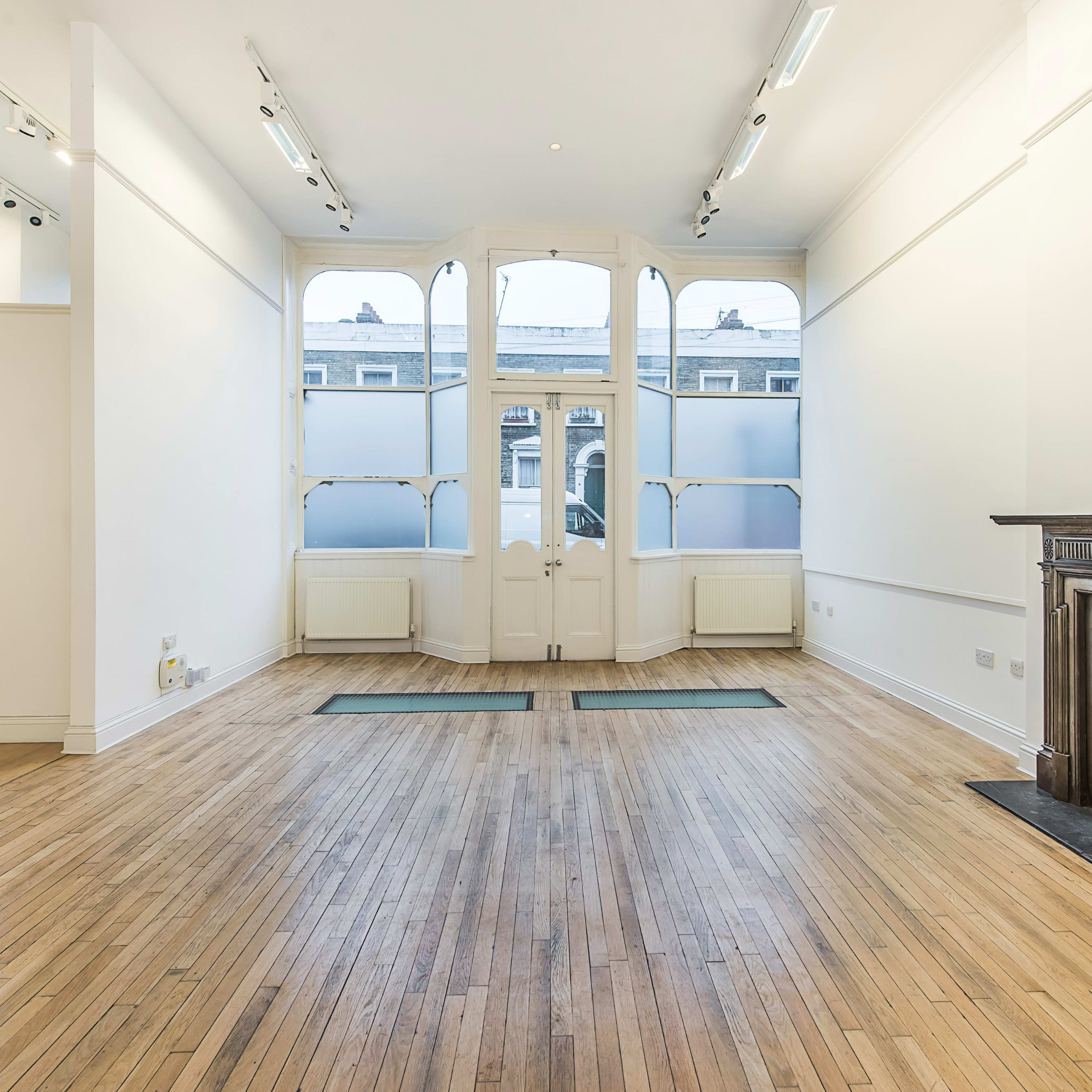 A spacious, empty room with wooden flooring, large windows, and an ornate fireplace against one wall.