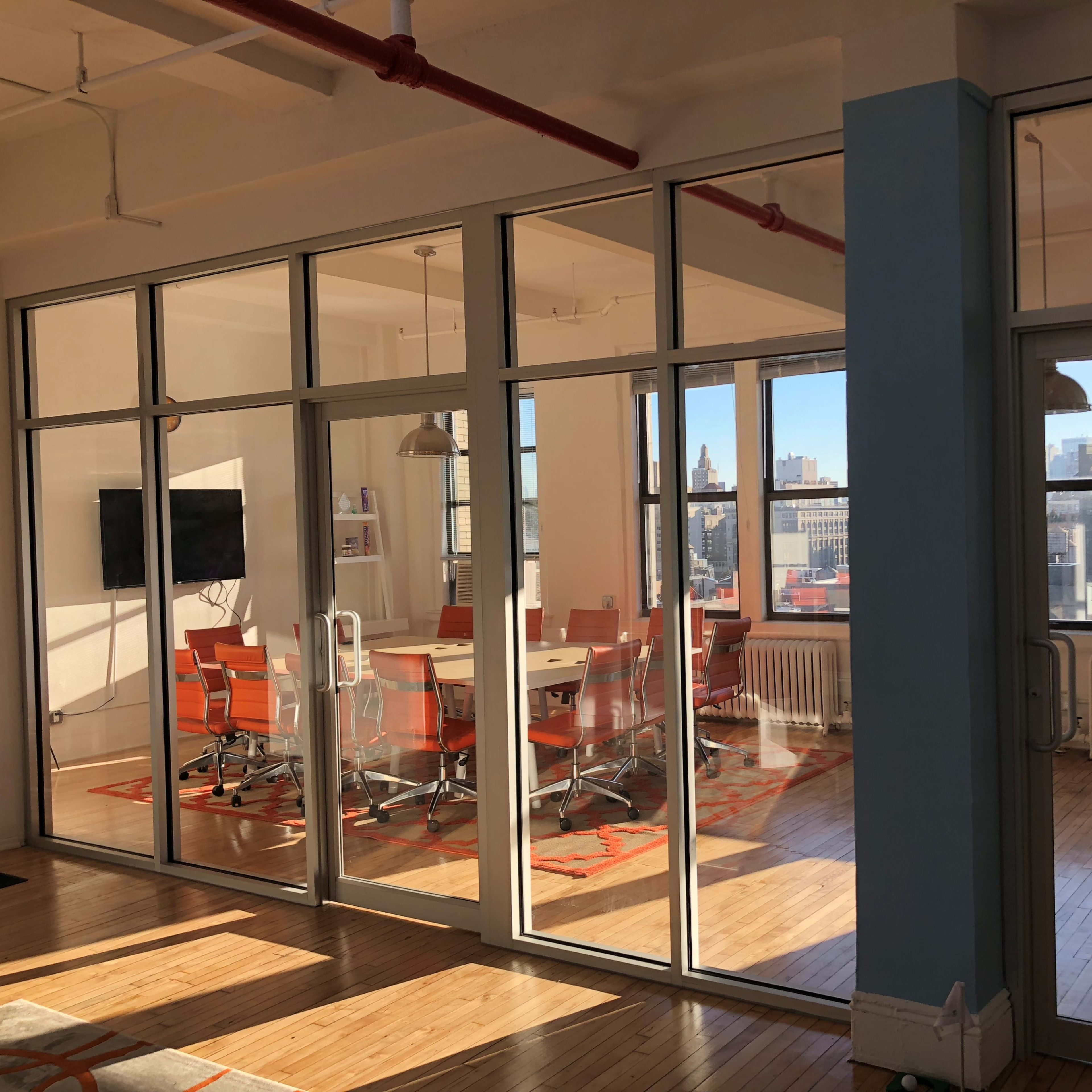 A modern conference room with glass walls, featuring a central table surrounded by orange chairs, with city skyline views through large windows.