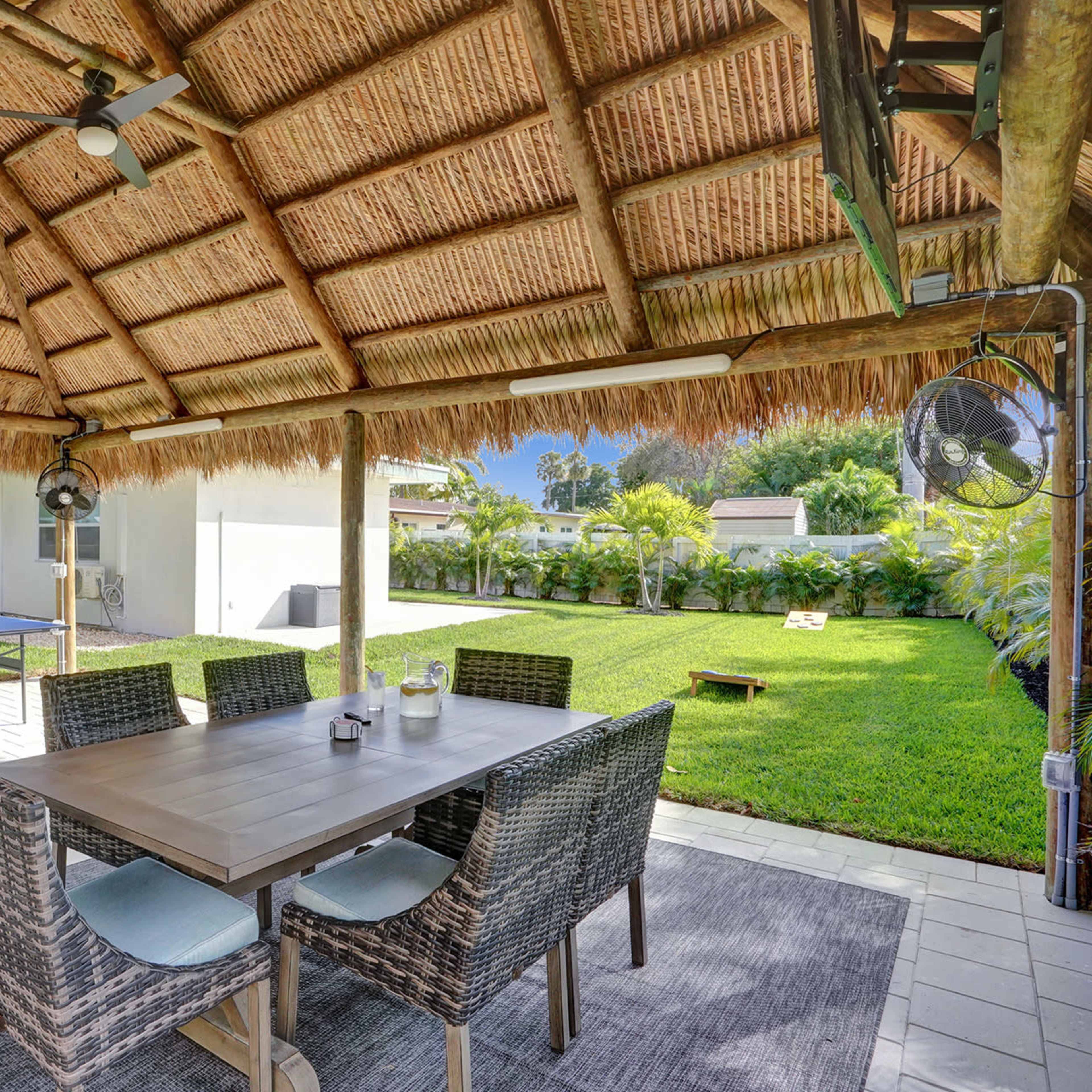 A shaded outdoor dining area with a table and chairs is set under a thatched roof, overlooking a grassy yard and a grill nearby.