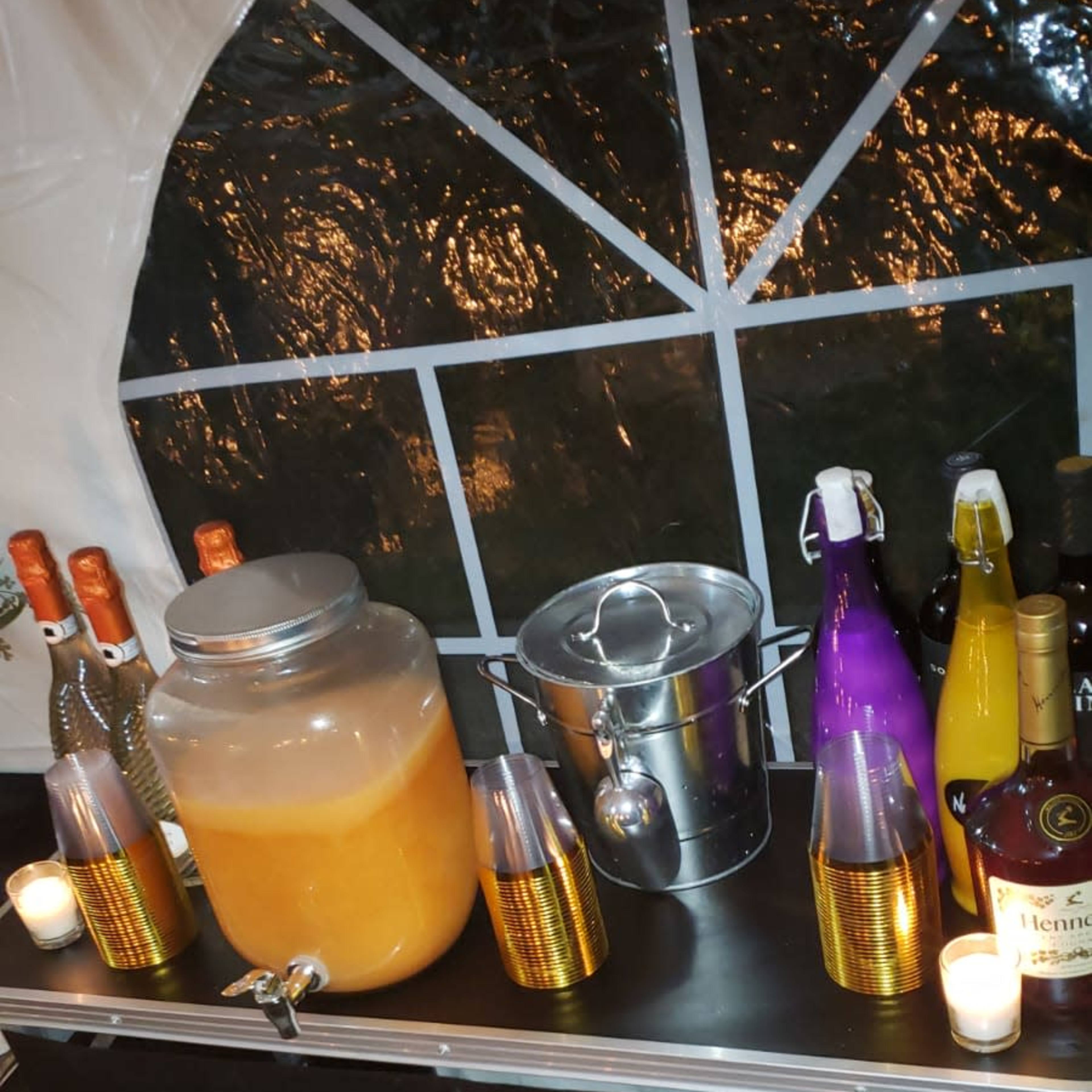 A bar setup inside a tent displays a large jar of orange beverage, an ice bucket, and various bottles of alcohol arranged alongside elegant glasses.