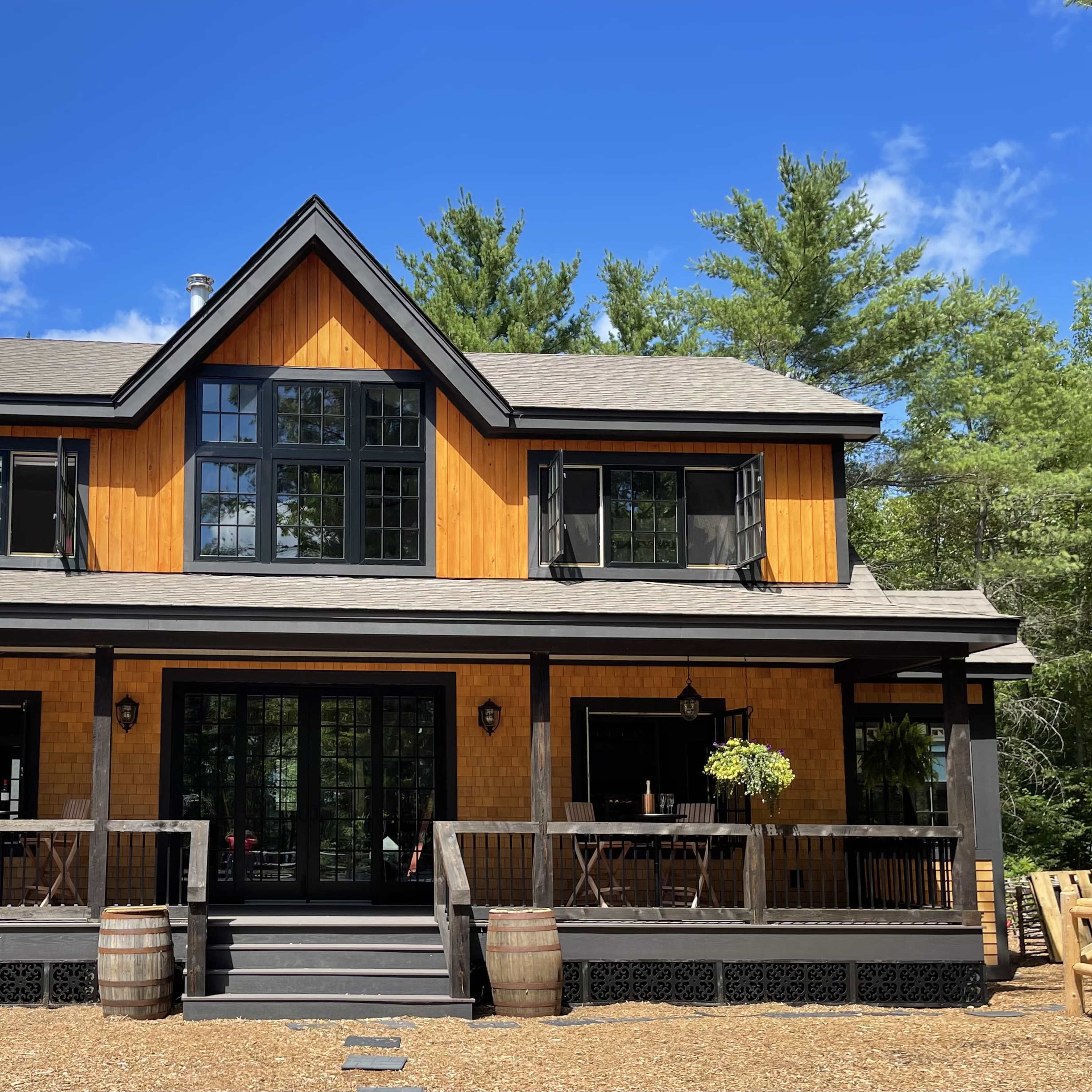 A two-story wooden house with large windows and a covered front porch, surrounded by trees and gravel.