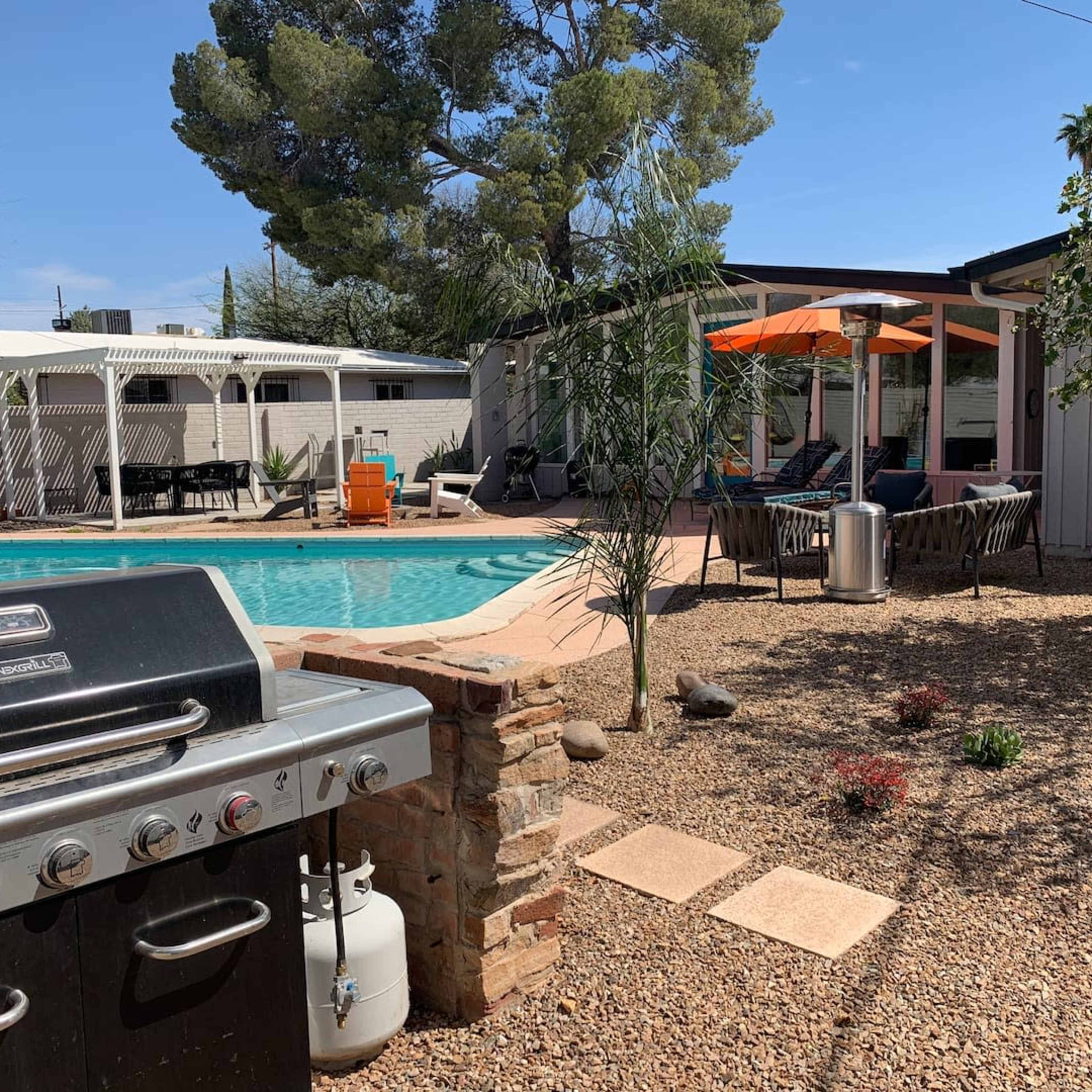 A backyard with a swimming pool, a barbecue grill, lounge chairs, and a covered seating area surrounded by gravel and greenery.
