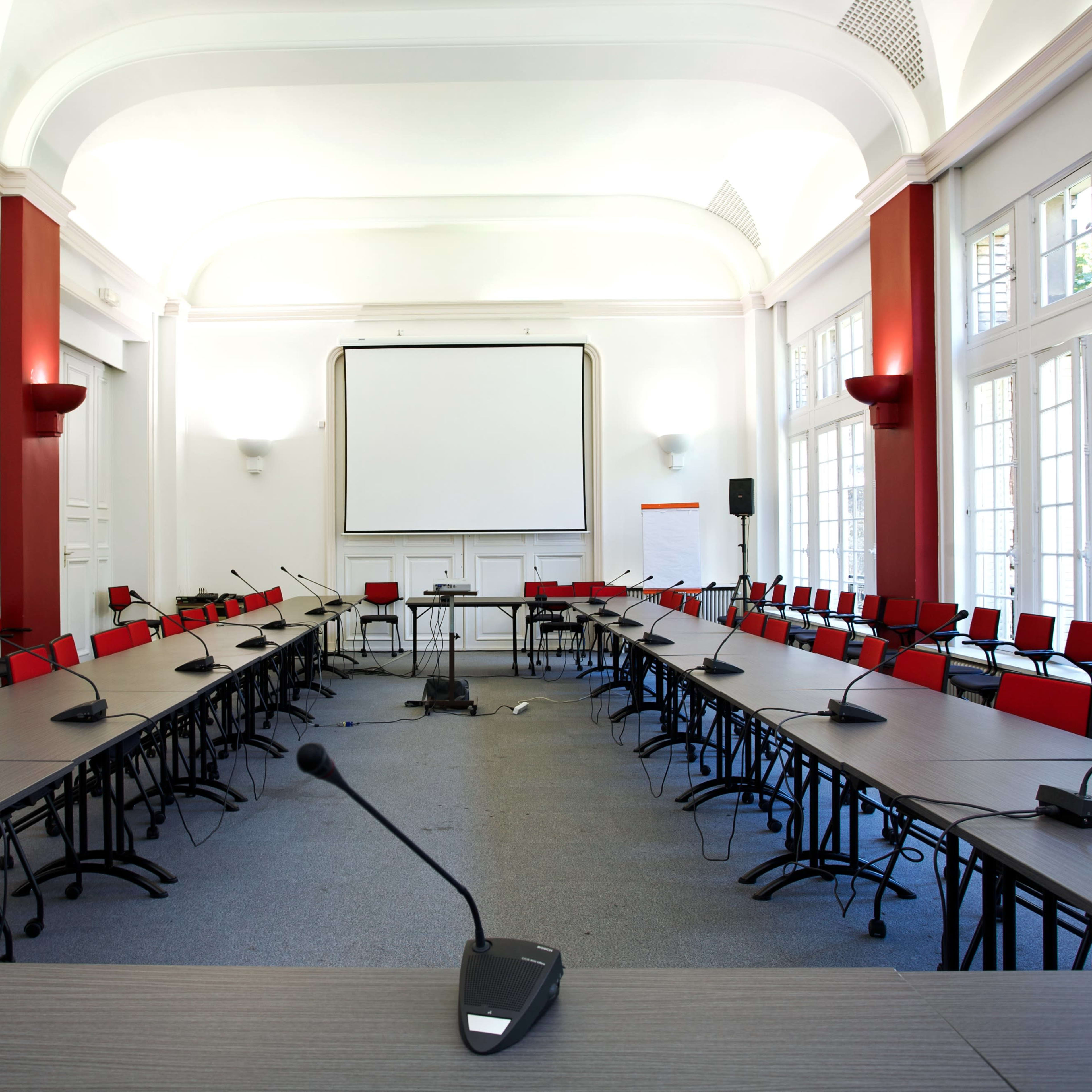 A spacious conference room arranged with multiple tables in a U-shape, surrounded by red chairs, and featuring a large screen at the front.