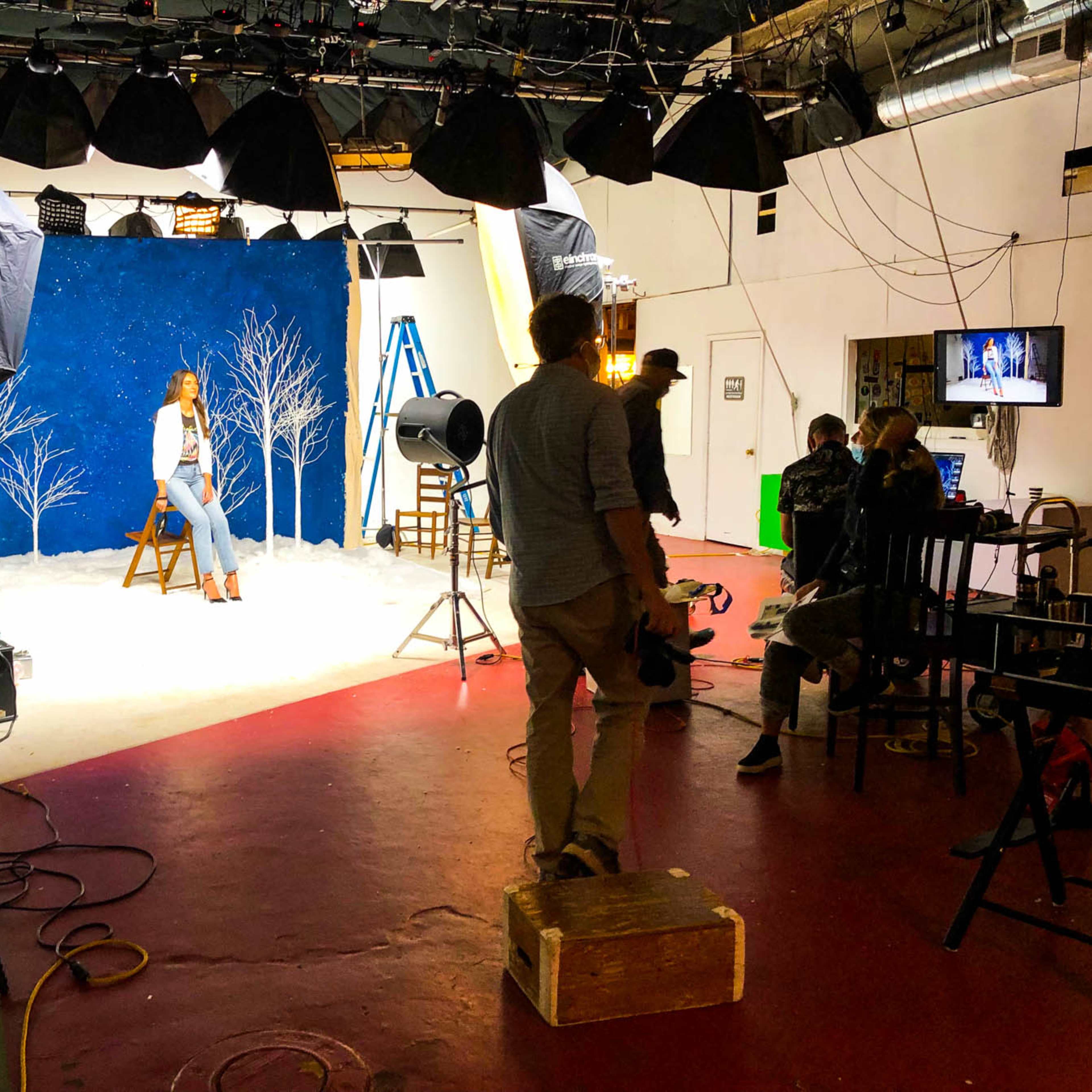 A photography studio is set up for a shoot, featuring a model on a stool in front of a blue backdrop with white tree decorations, while crew members and equipment are arranged around the scene.
