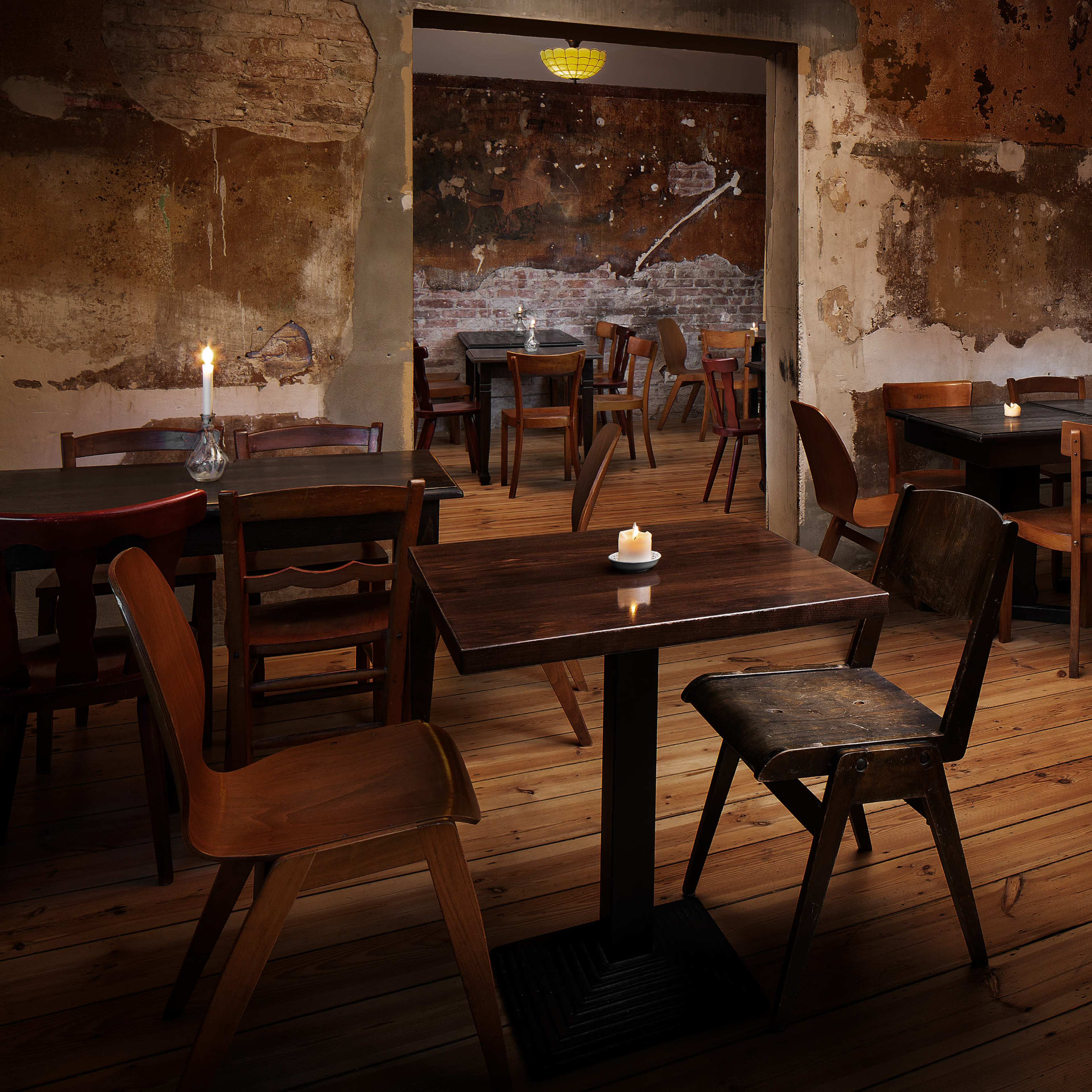 A rustic interior of a casual dining space with wooden tables and chairs arranged around a dimly lit room featuring exposed brick and peeling walls.