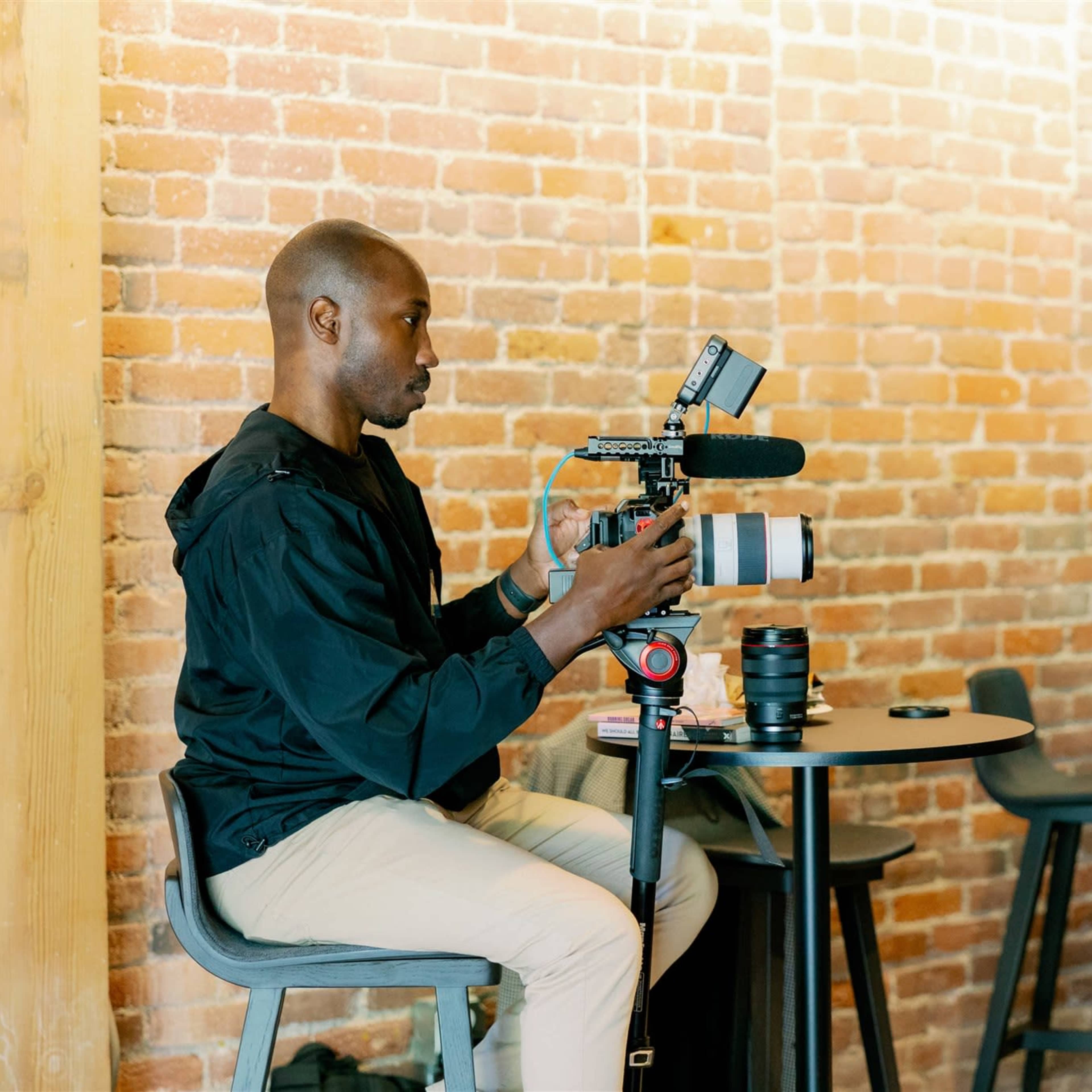 Two individuals operate cameras in a space with exposed brick walls, while a third person is seated at a table with filming equipment.