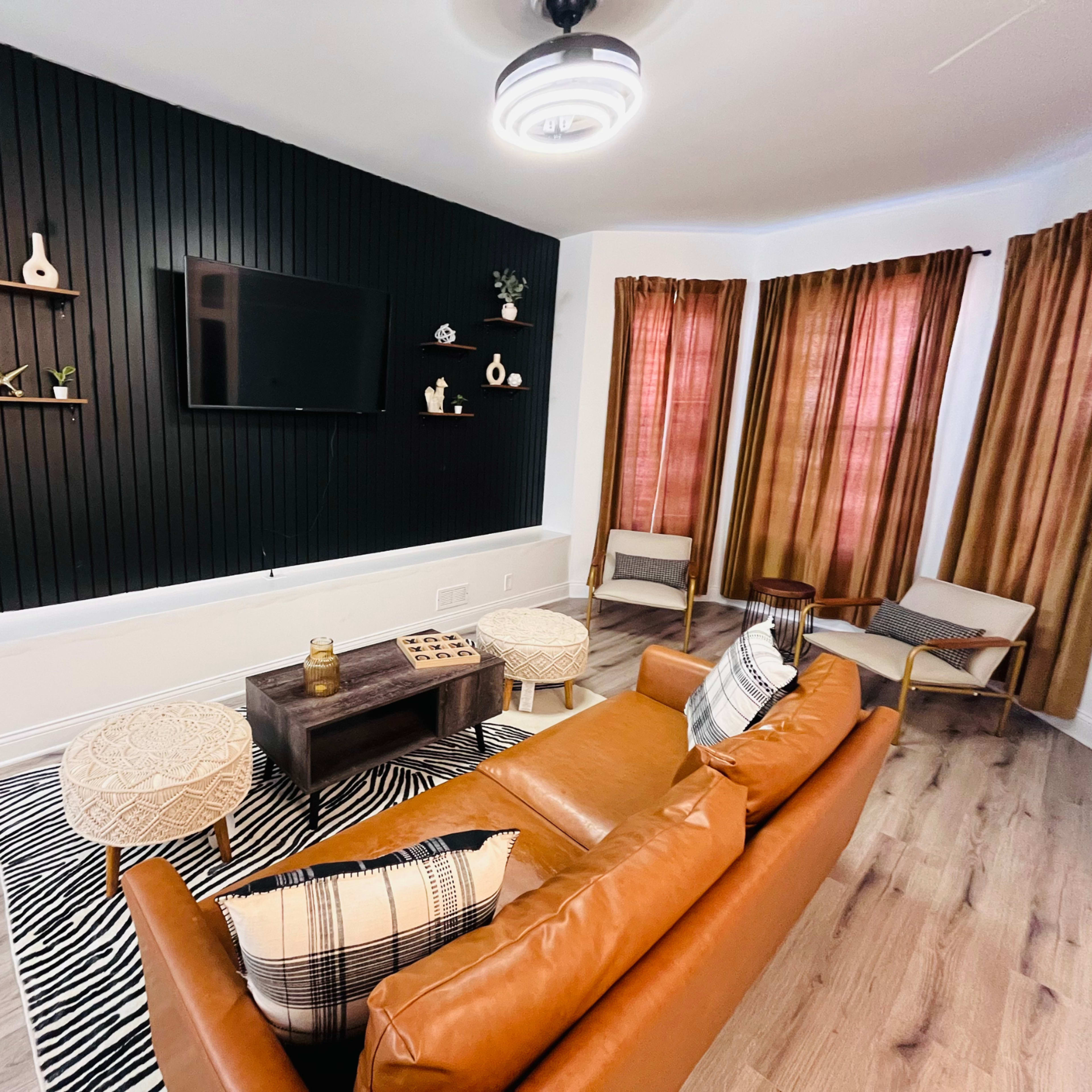 A modern living room with a brown leather sofa, a flat-screen TV mounted on a black paneled wall, and large windows draped with orange curtains.
