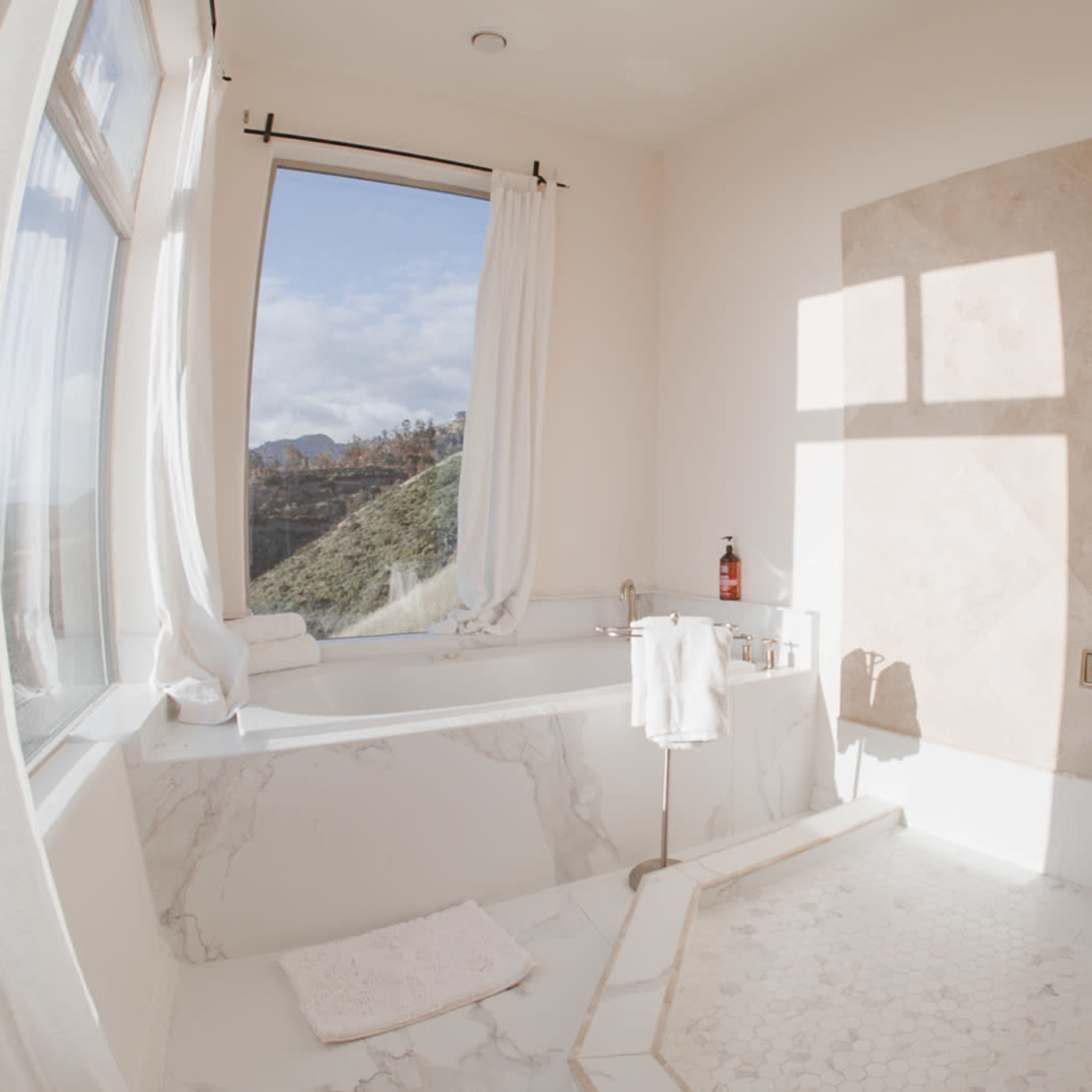 A modern bathroom features a bathtub beside a large window with a scenic view, and marble-tiled walls and floor.