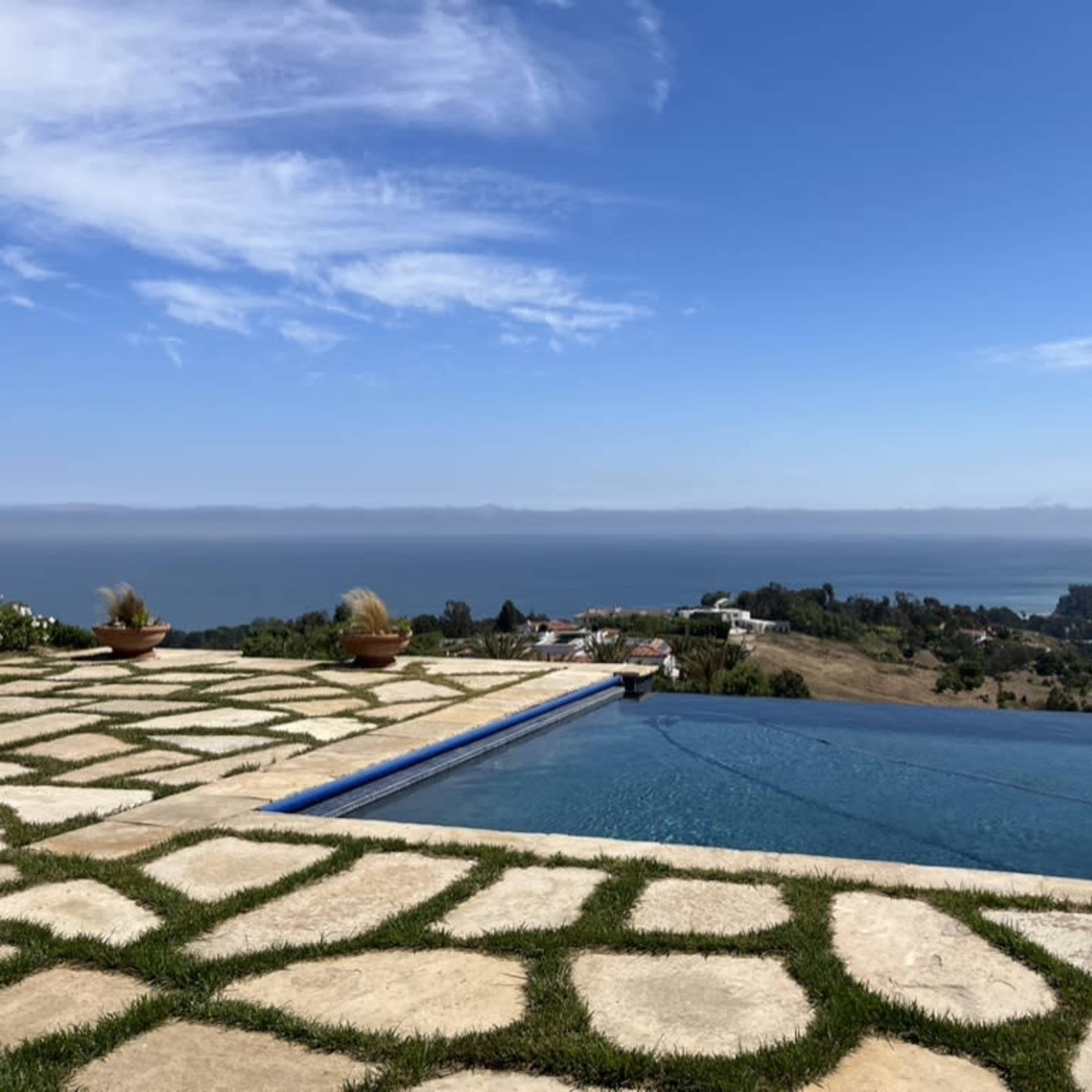 A swimming pool with an infinity edge overlooking a coastal landscape and the ocean under a clear blue sky.
