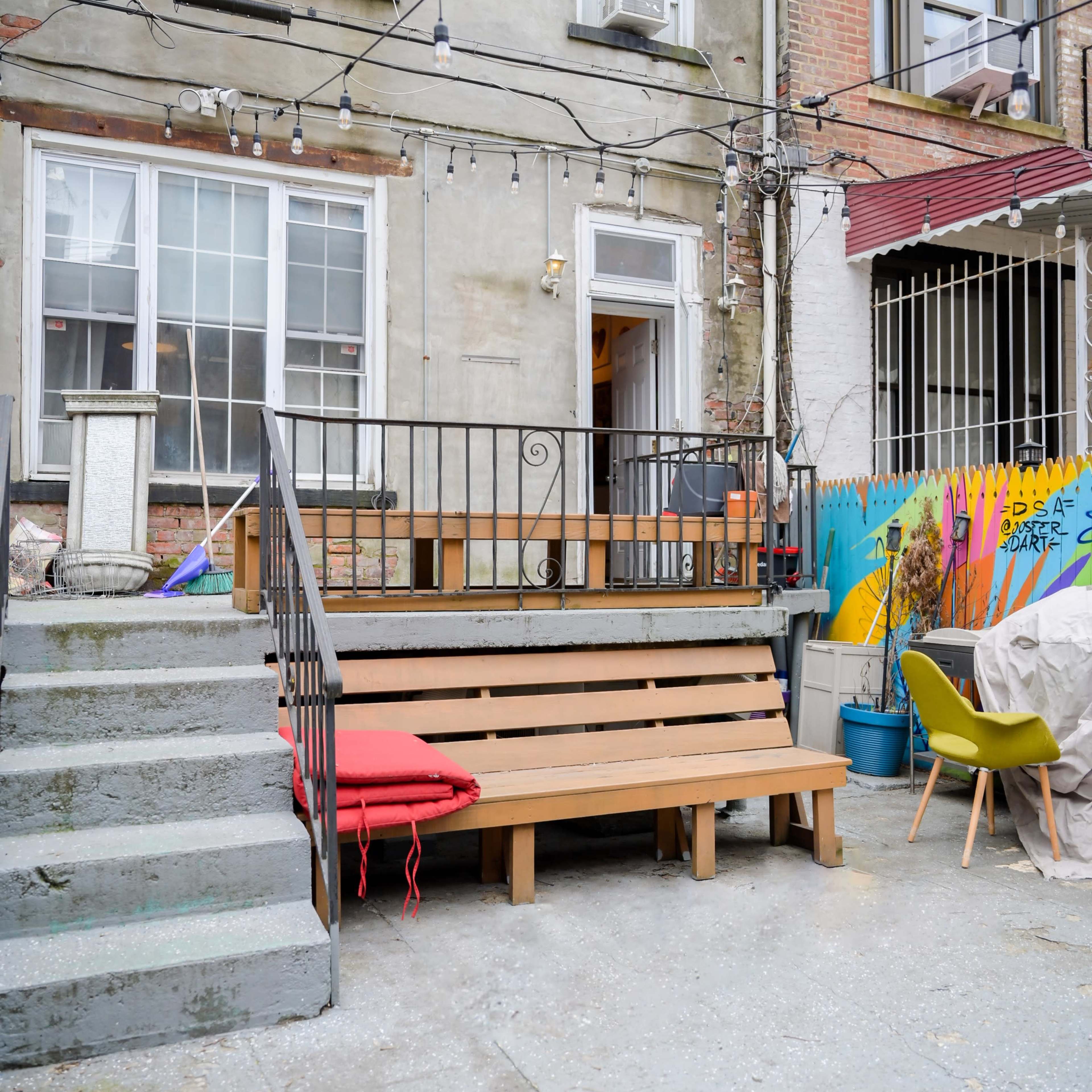 A small outdoor seating area with wooden benches and steps leading up to a building, featuring string lights and colorful wall art in the background.