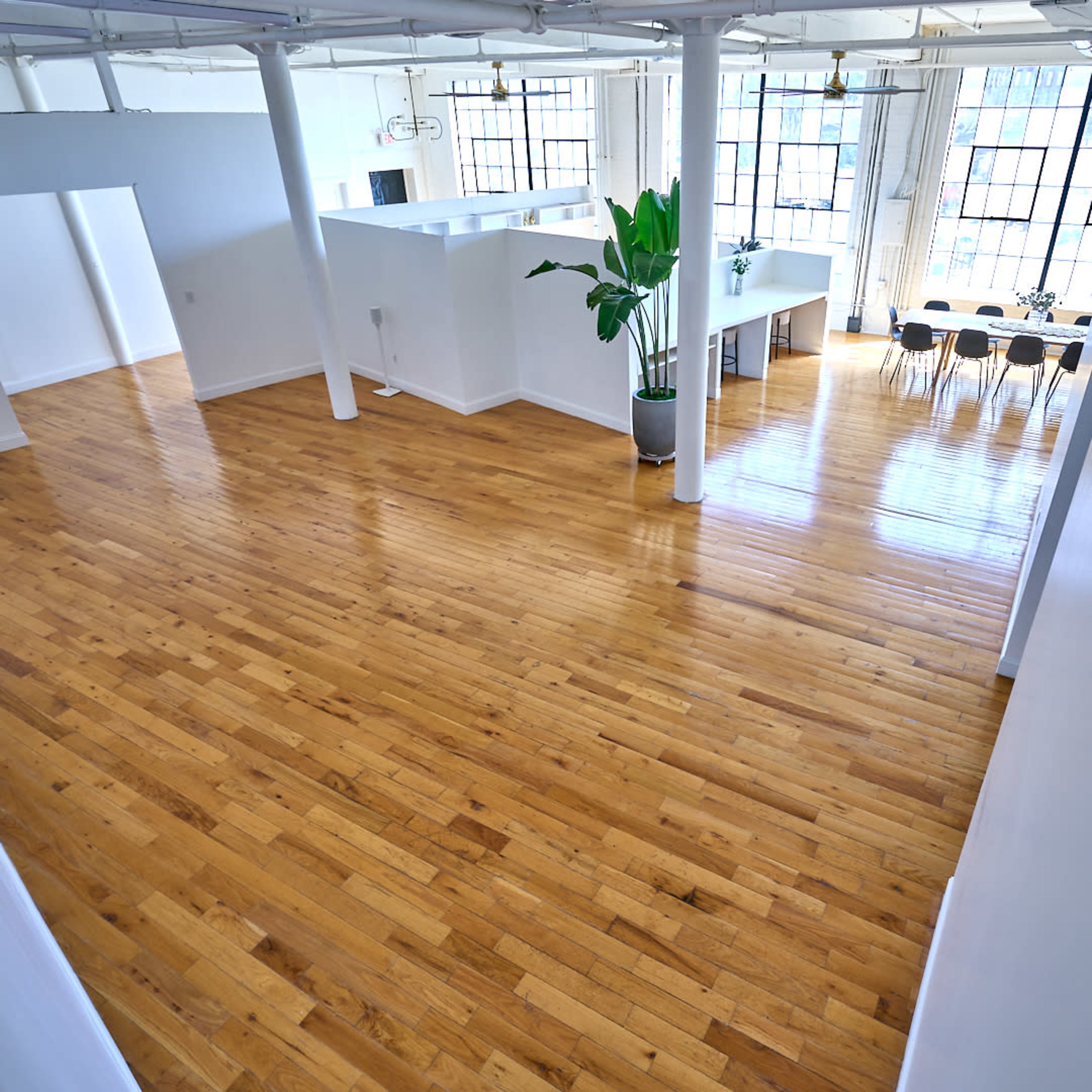 A spacious, well-lit room with polished wooden floors and large windows, featuring a dining area and a potted plant.