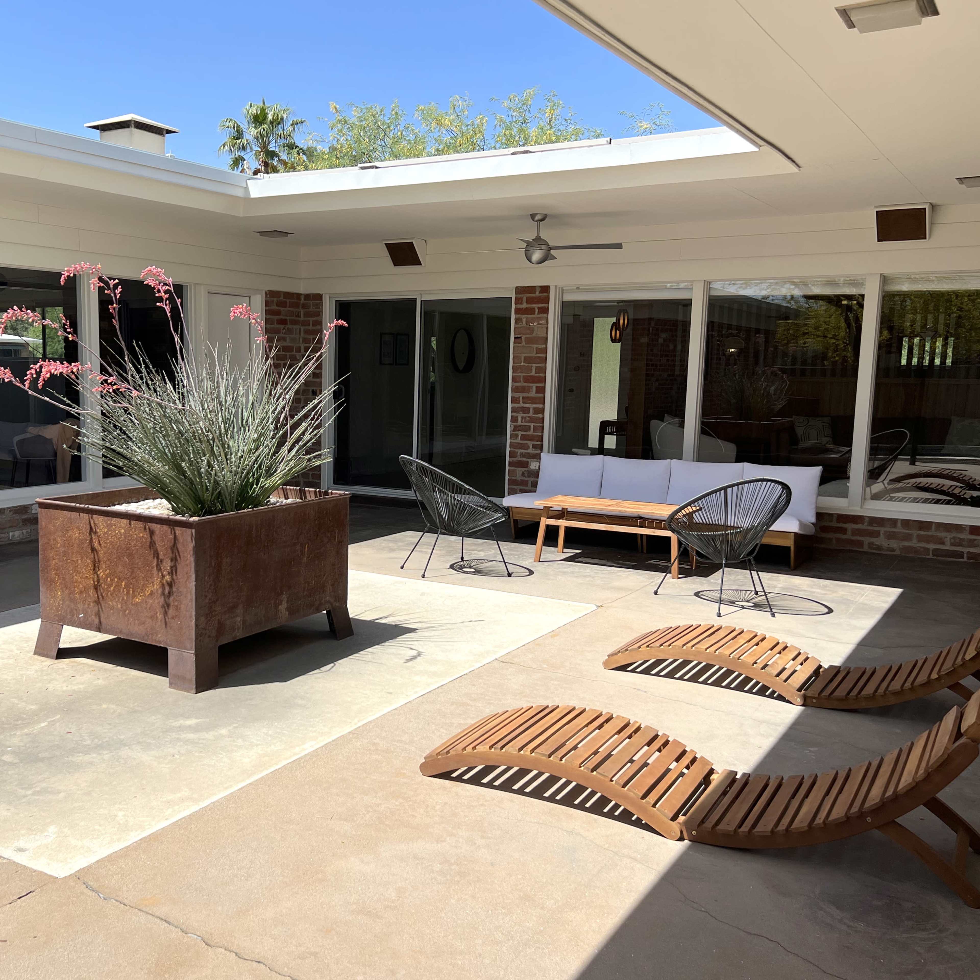 A modern outdoor patio featuring a large planter, two lounge chairs, a wooden coffee table, and several large glass doors leading into an interior space.