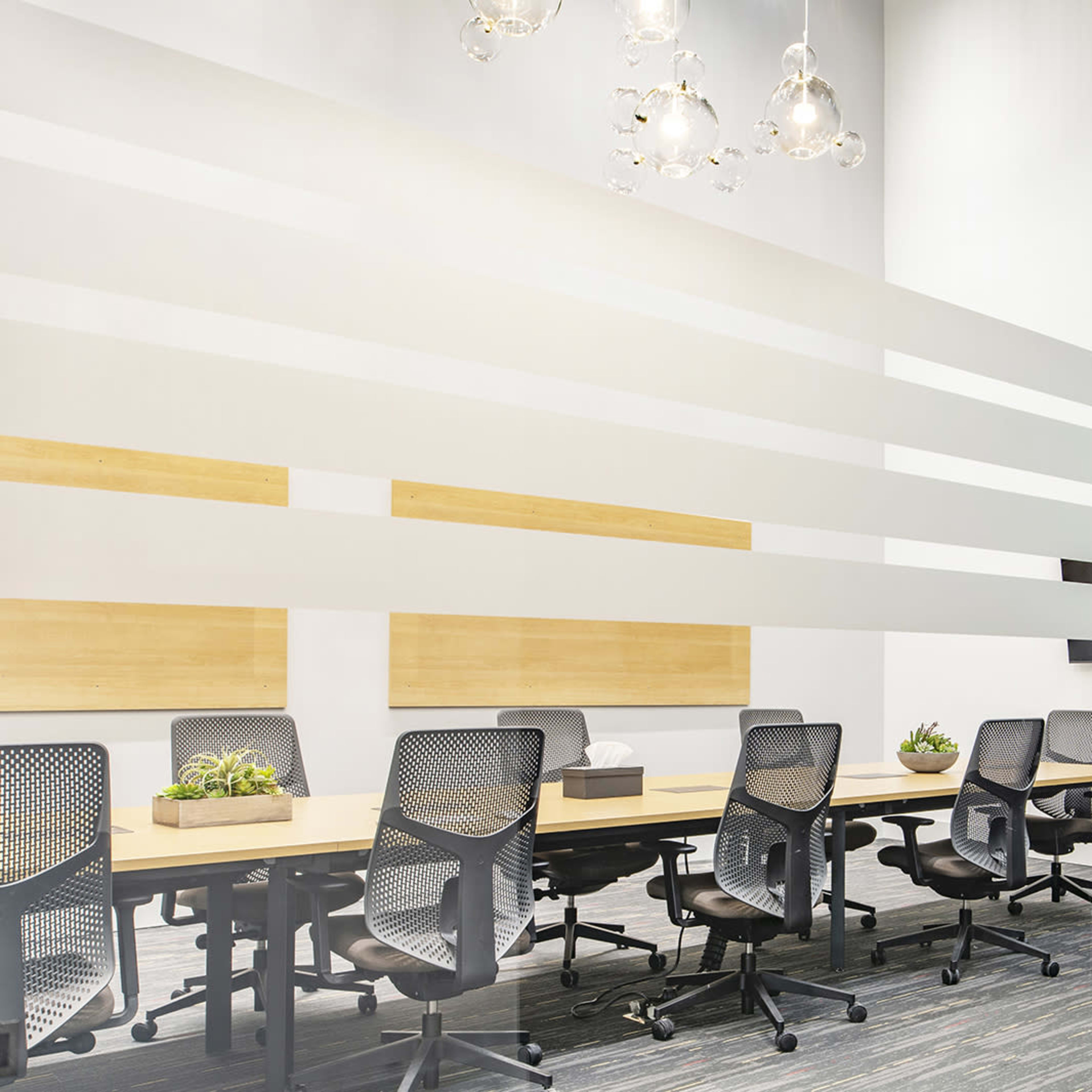 A modern conference room features a long table surrounded by several chairs, with large glass walls and decorative planters.