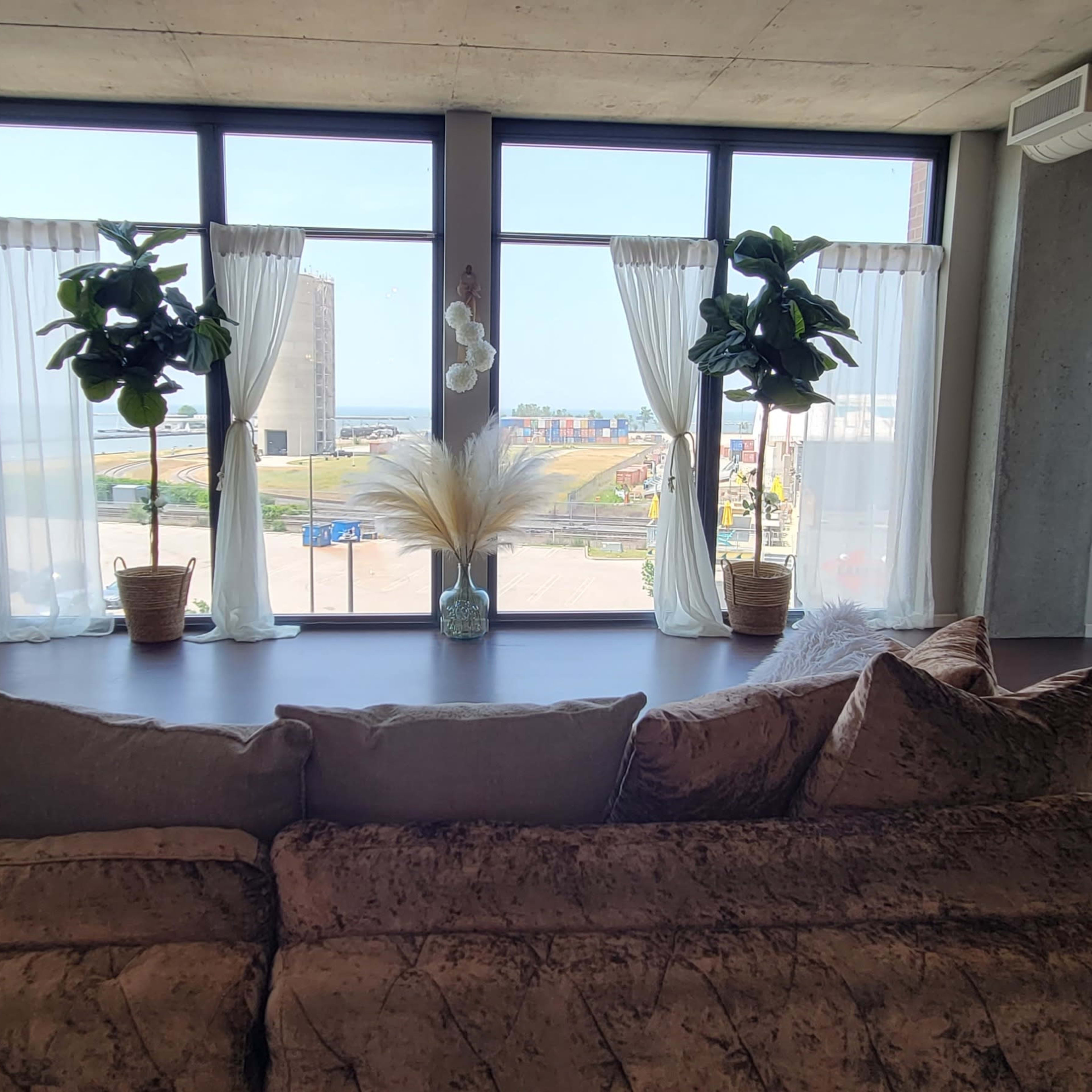 A modern living room with a large window revealing a view of an outdoor area, featuring a plush sofa, decorative plants, and sheer curtains.
