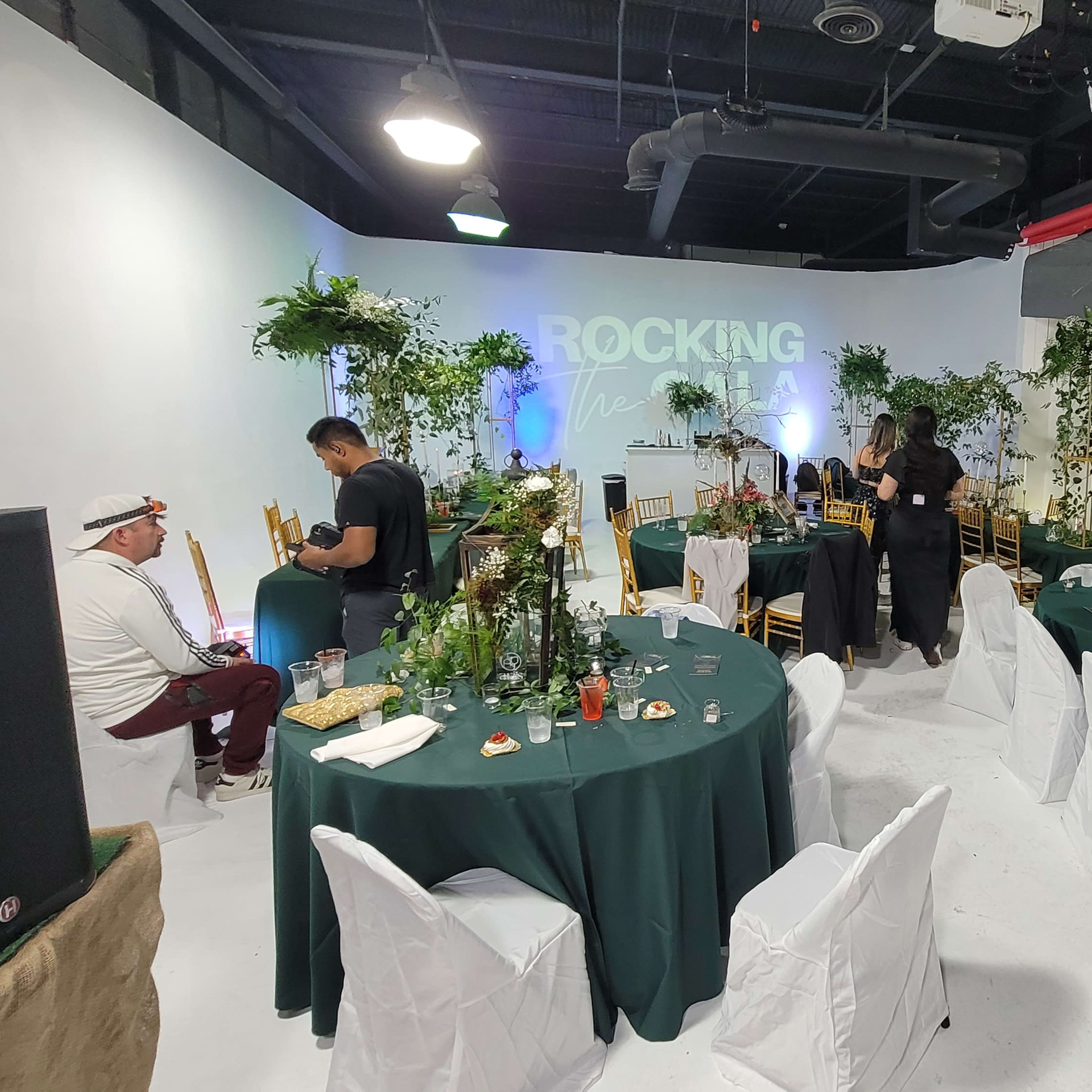 A spacious event venue features round tables set with green tablecloths, surrounded by guests mingling among decorative plants and a backdrop that reads "ROCKING THE V".