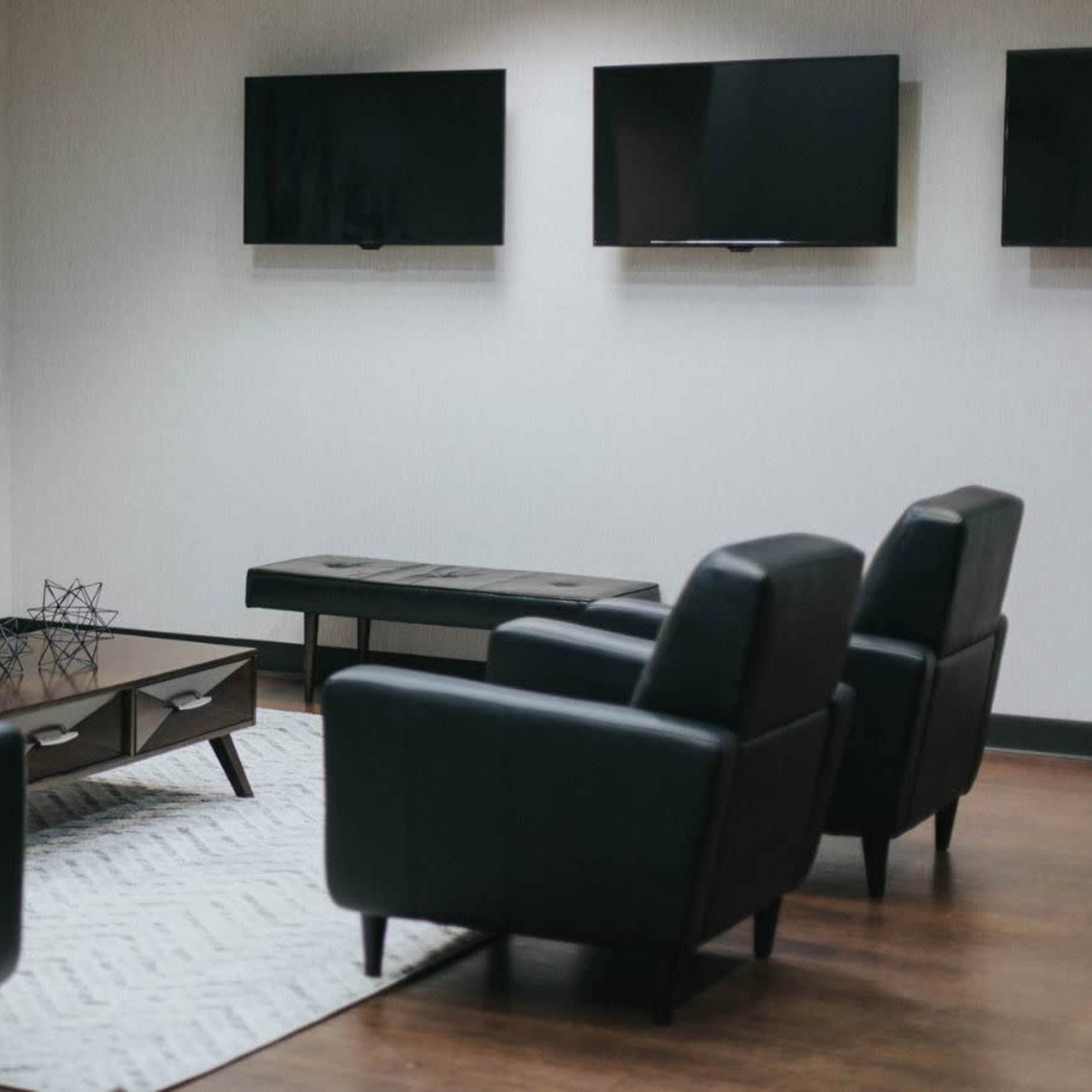 A minimalist lounge area with two black couches, four armchairs, a coffee table, and two mounted TVs on the wall.