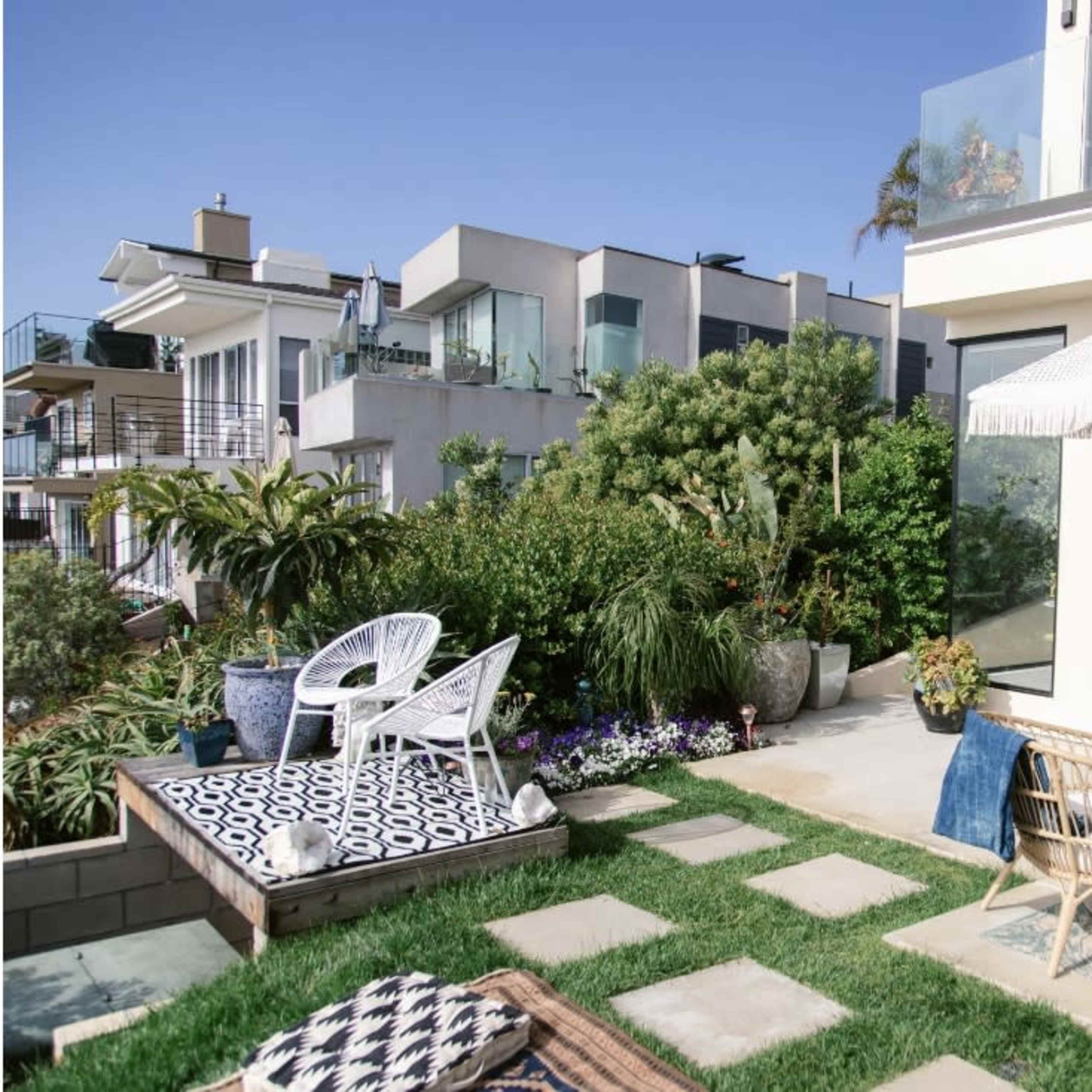 A landscaped patio area features white outdoor chairs, a patterned rug, and a large umbrella beside a modern home.