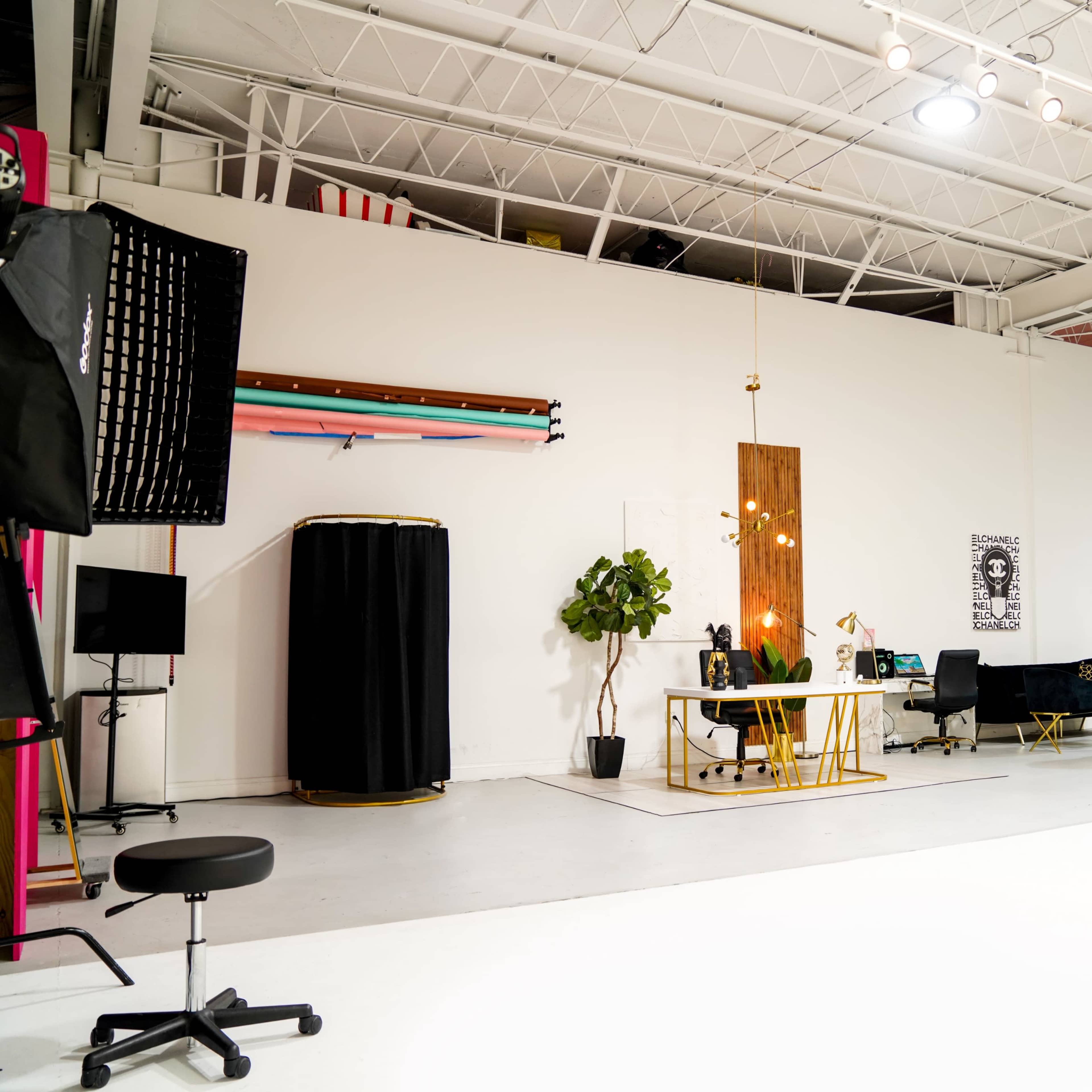 A spacious photography studio with a reception area featuring a desk, plants, and stylish black furnishings, along with equipment and backdrops arranged along the walls.