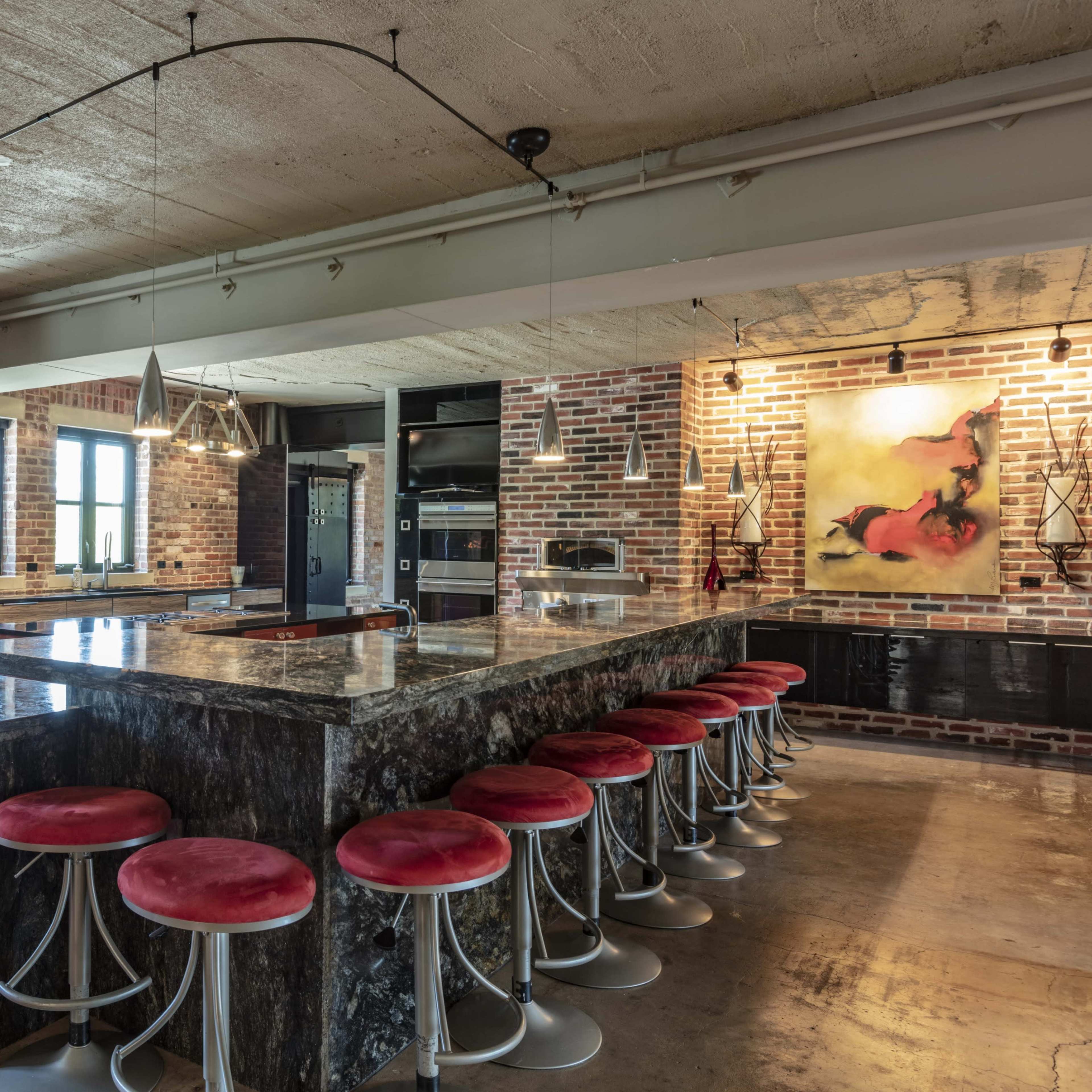 A modern kitchen features a large island with a dark countertop and red bar stools, complemented by exposed brick walls and industrial-style lighting.