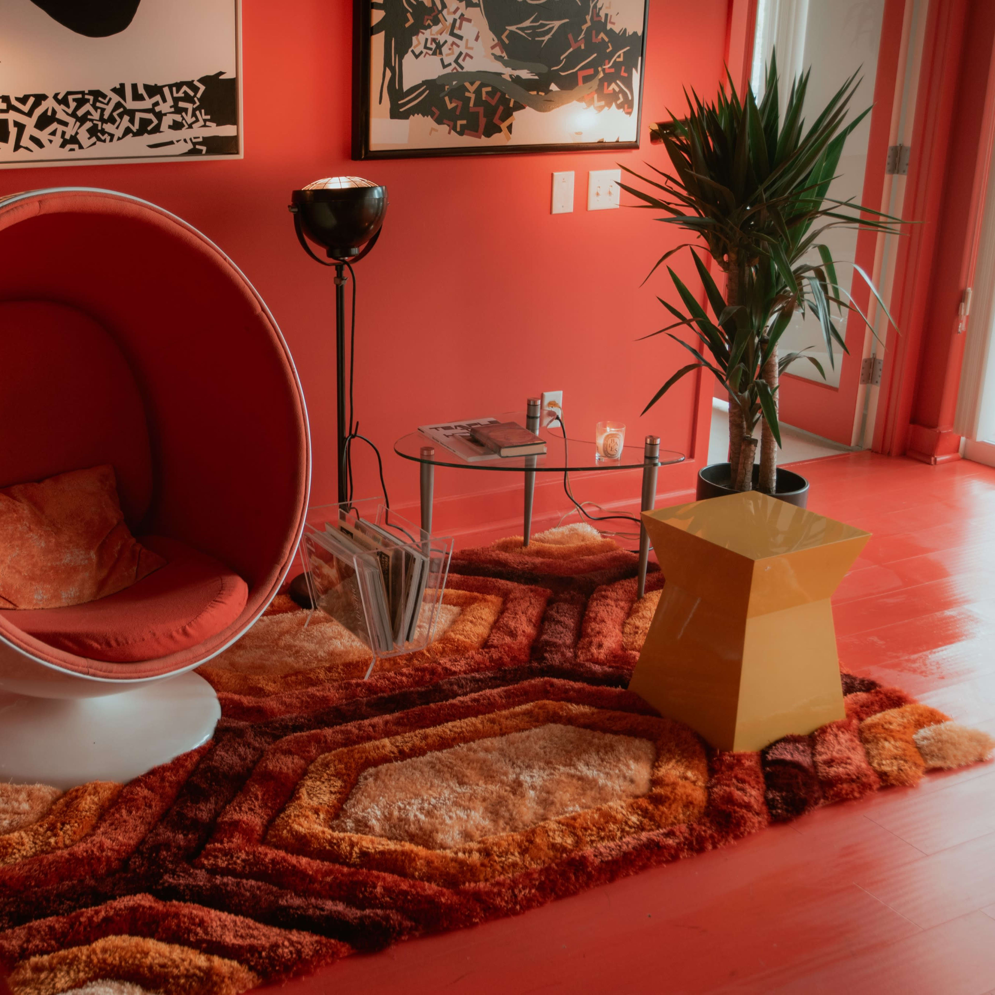 A modern red-themed living space features a round, white chair, a black lamp, a green plant, and a geometric yellow table on a colorful, textured rug.