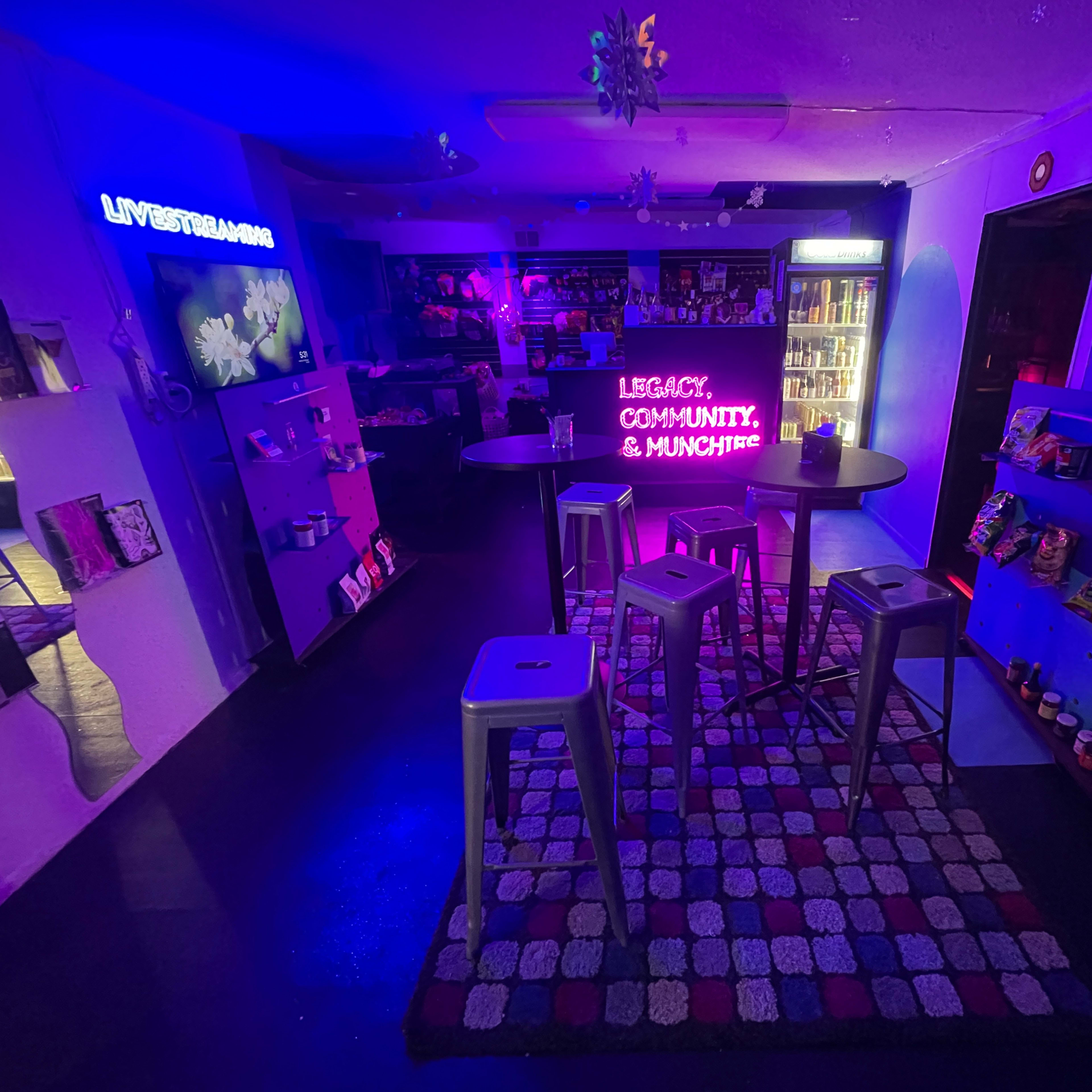 A colorful bar area with modern furniture, featuring neon signs and shelves filled with snacks.