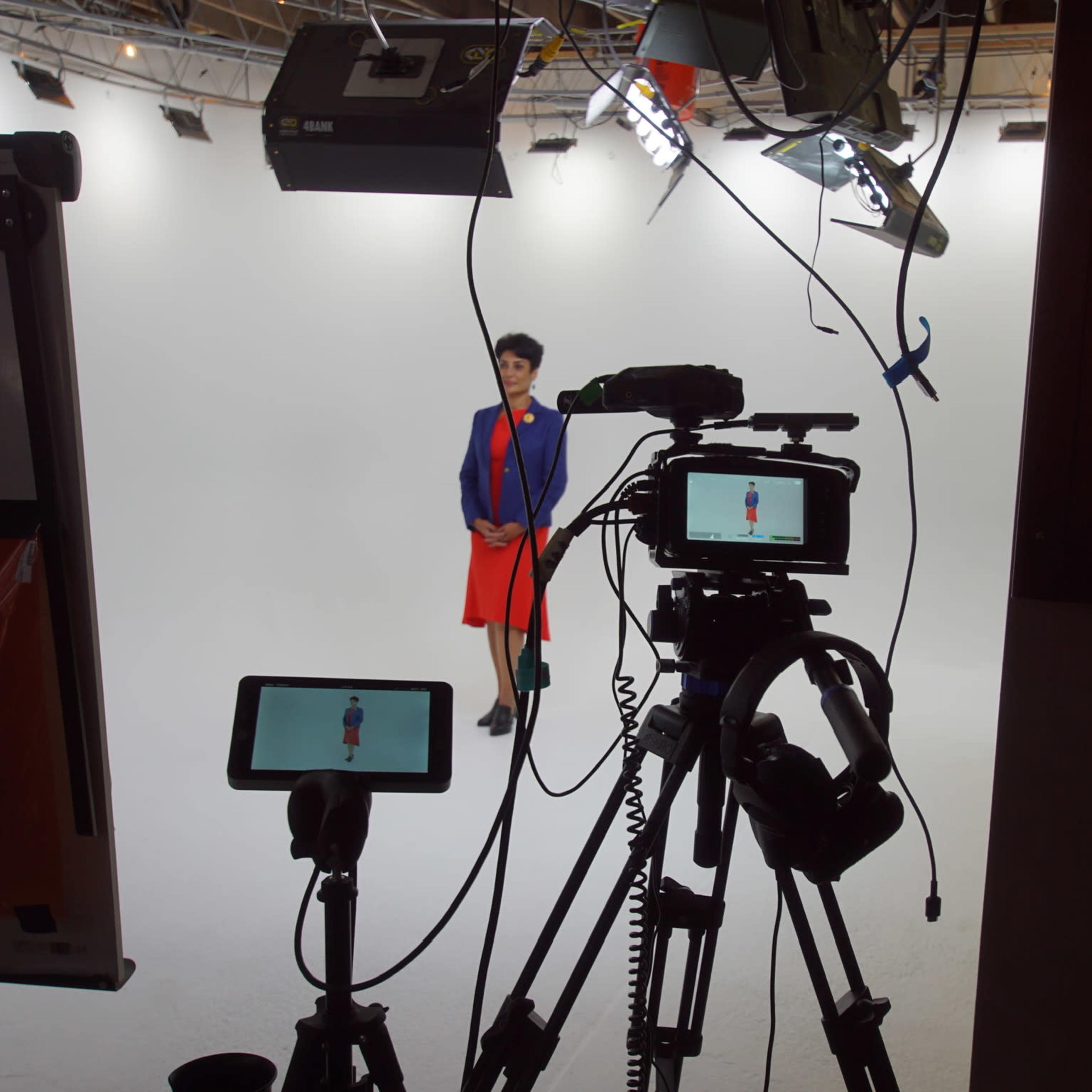 A woman in a blue jacket and red skirt stands against a white backdrop, surrounded by video cameras and lighting equipment.