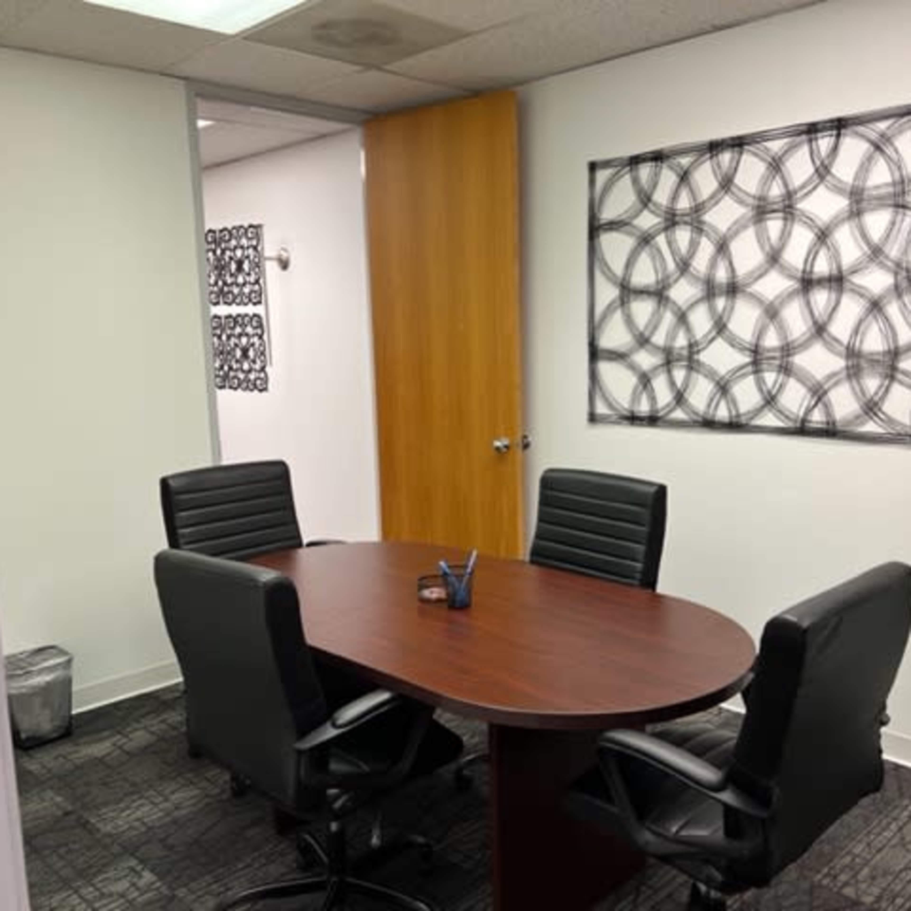 A conference room is furnished with a round wooden table and four black leather chairs, featuring a framed artwork on the wall.