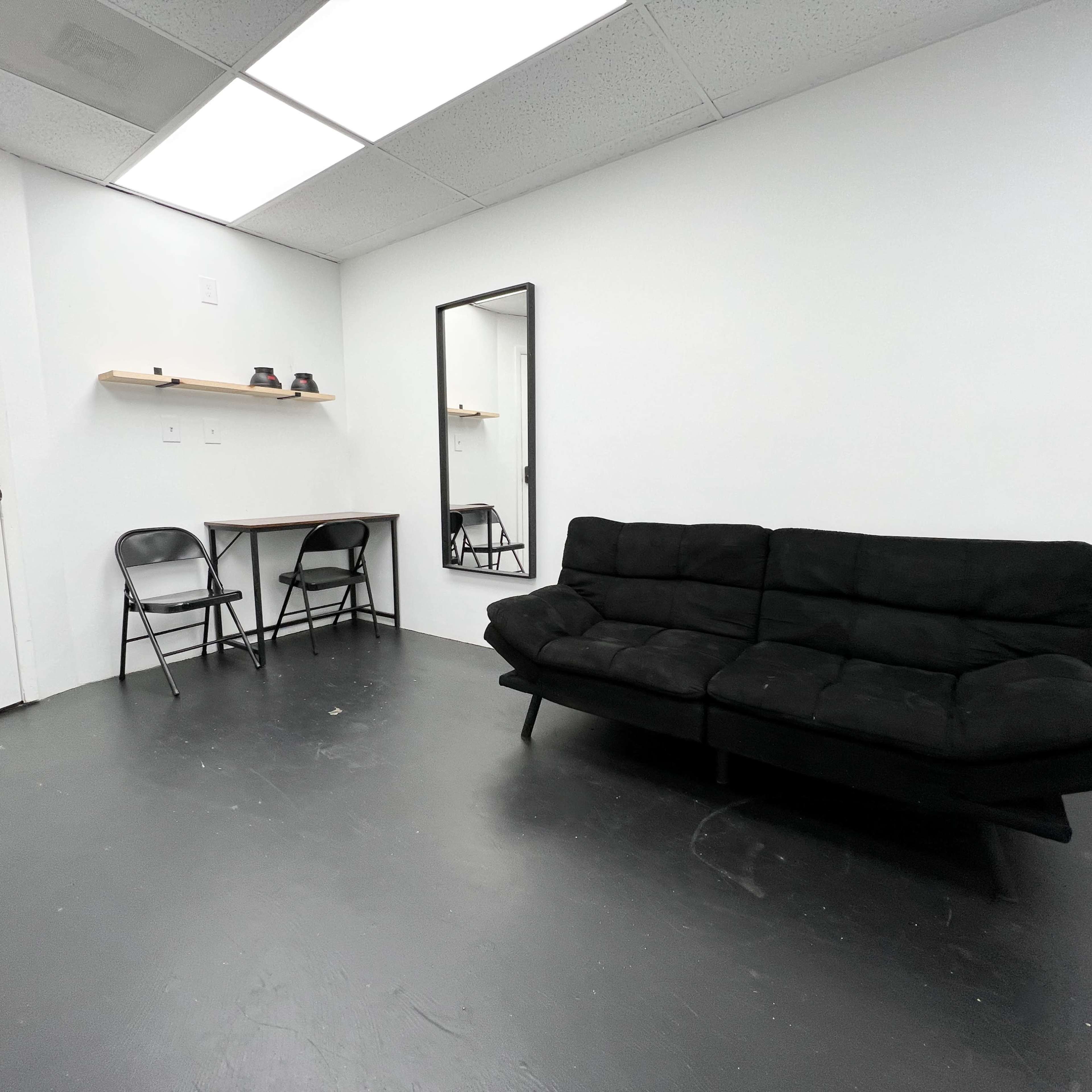 A minimalistic room with a black sofa, a small table with two chairs, and a mirror on a white wall.